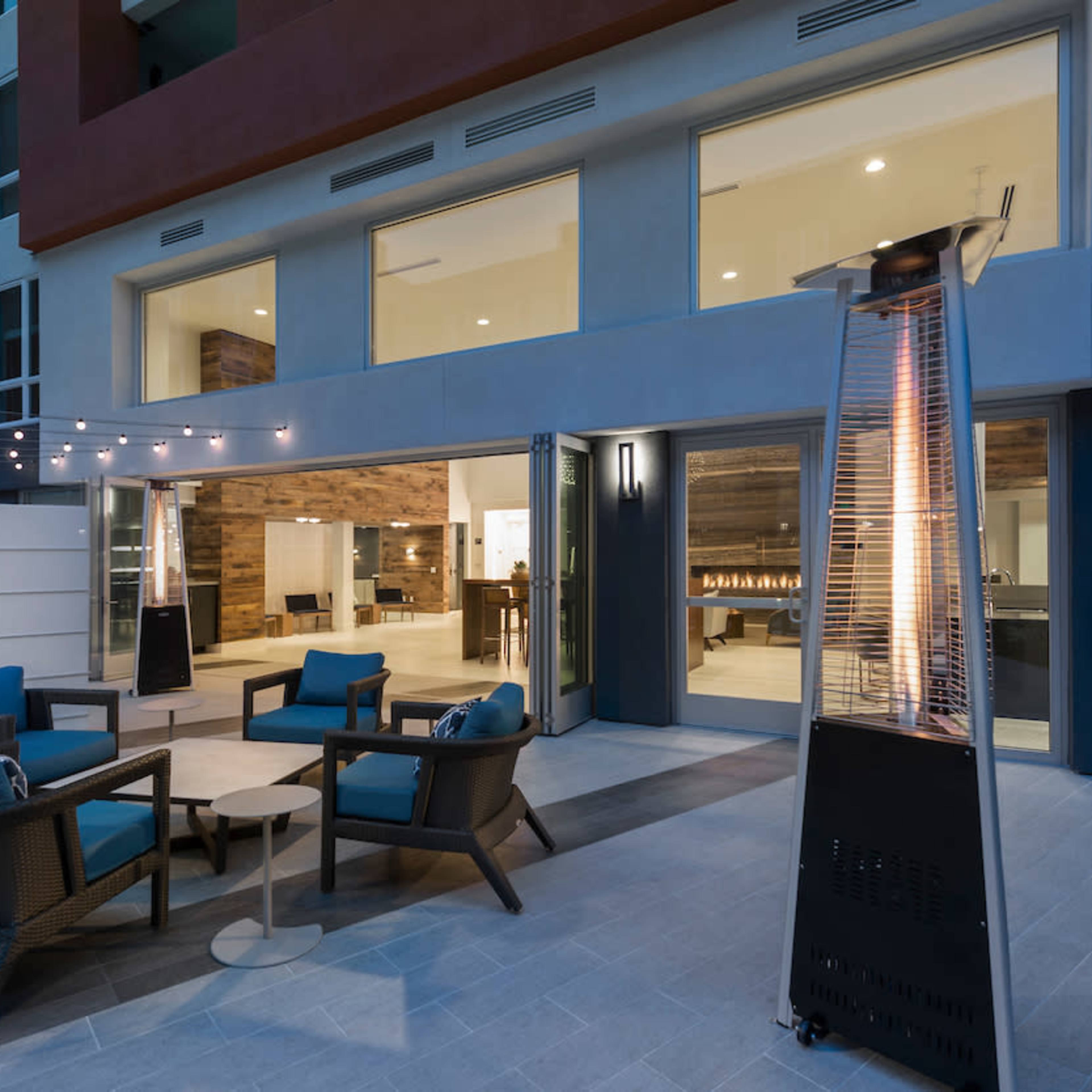 A modern outdoor patio with seating, a gas heater, and string lights, adjacent to a large glass windowed area of a building.