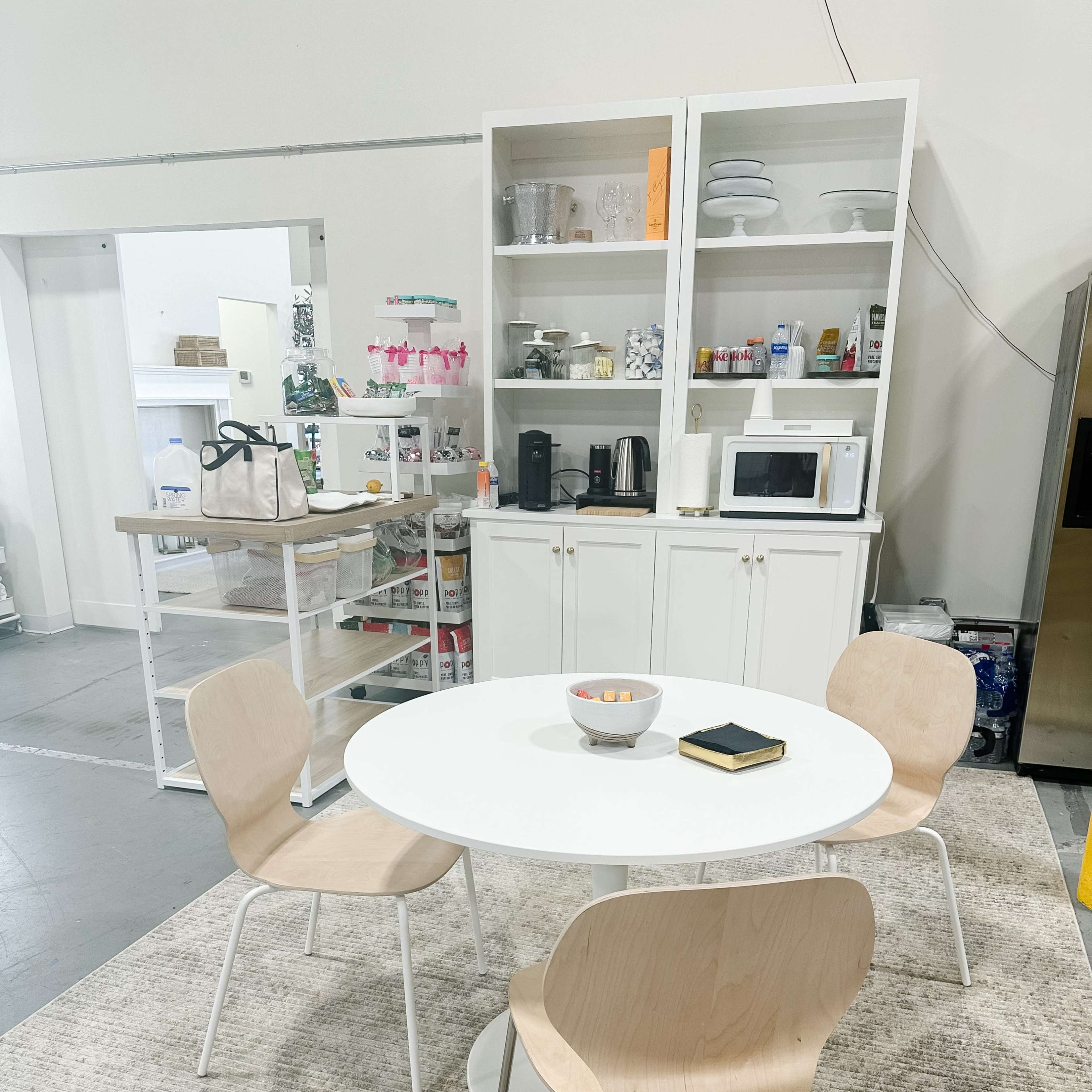 A modern kitchen area with a round white table, three wooden chairs, a bowl of fruit on the table, and a countertop with kitchen appliances and dishes displayed in a glass-front cabinet.