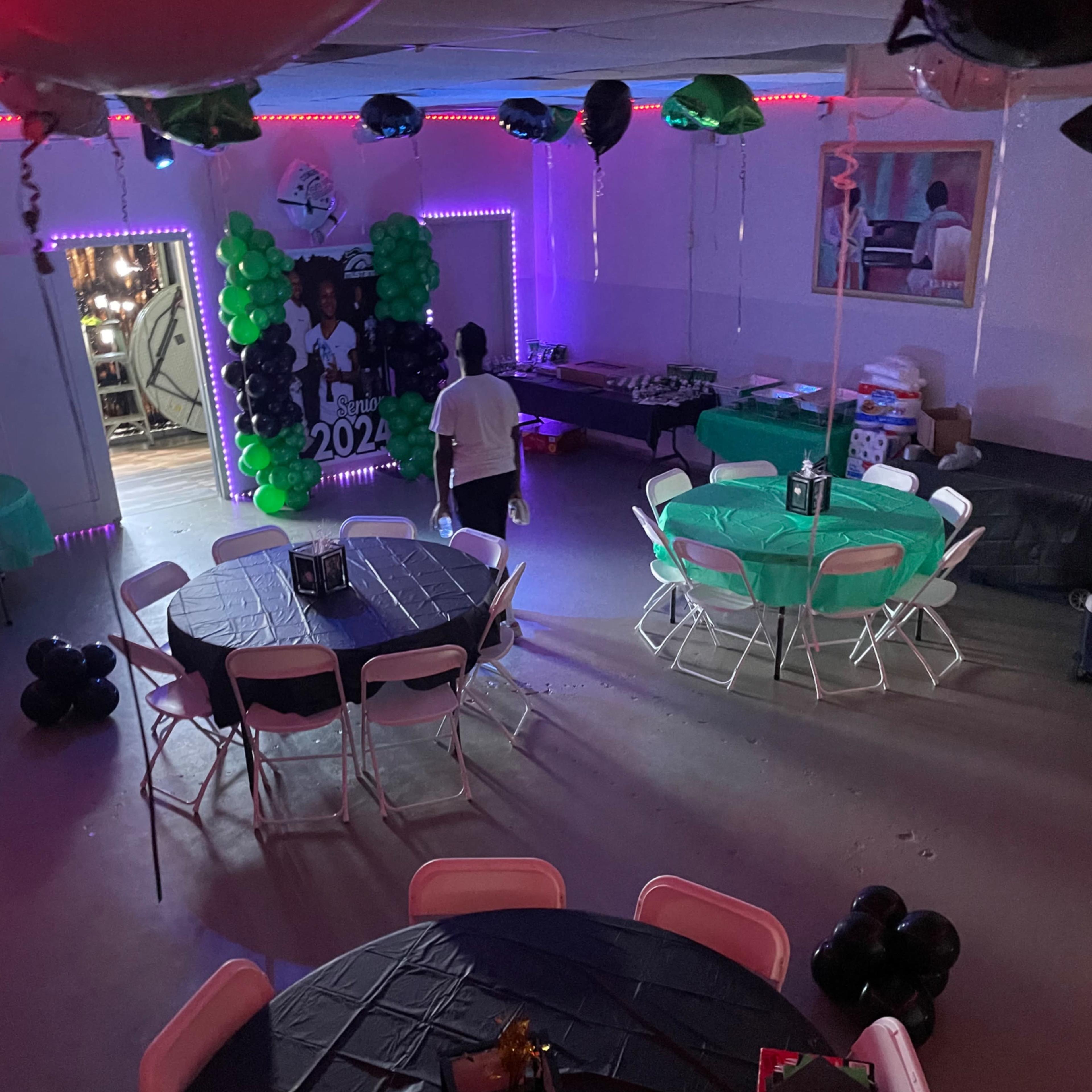 A decorated party space with round tables covered in black tablecloths, green balloons, and a backdrop celebrating a graduation in 2024.