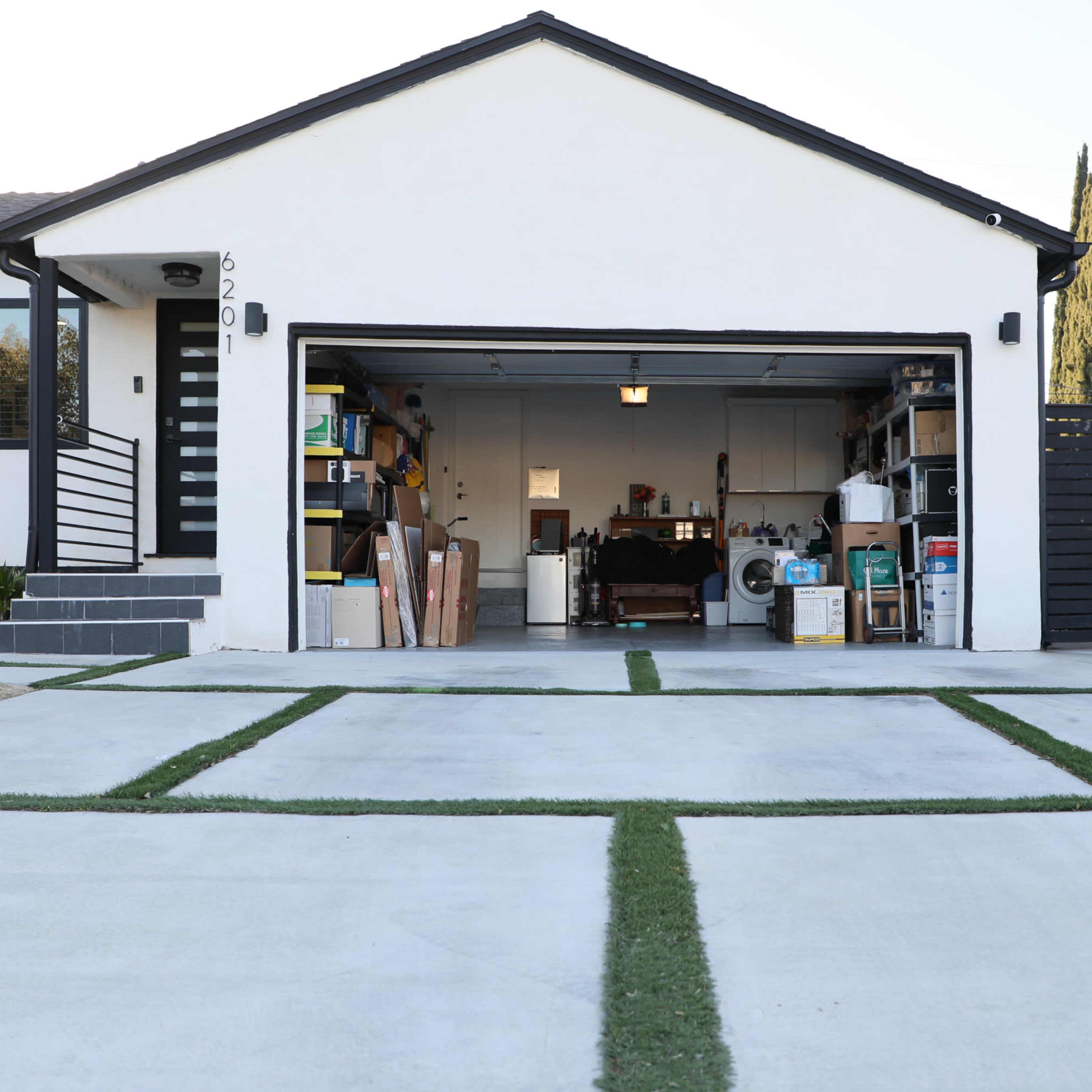 A garage with an open door reveals organized shelves and items, accompanied by a clean driveway featuring grass strips.