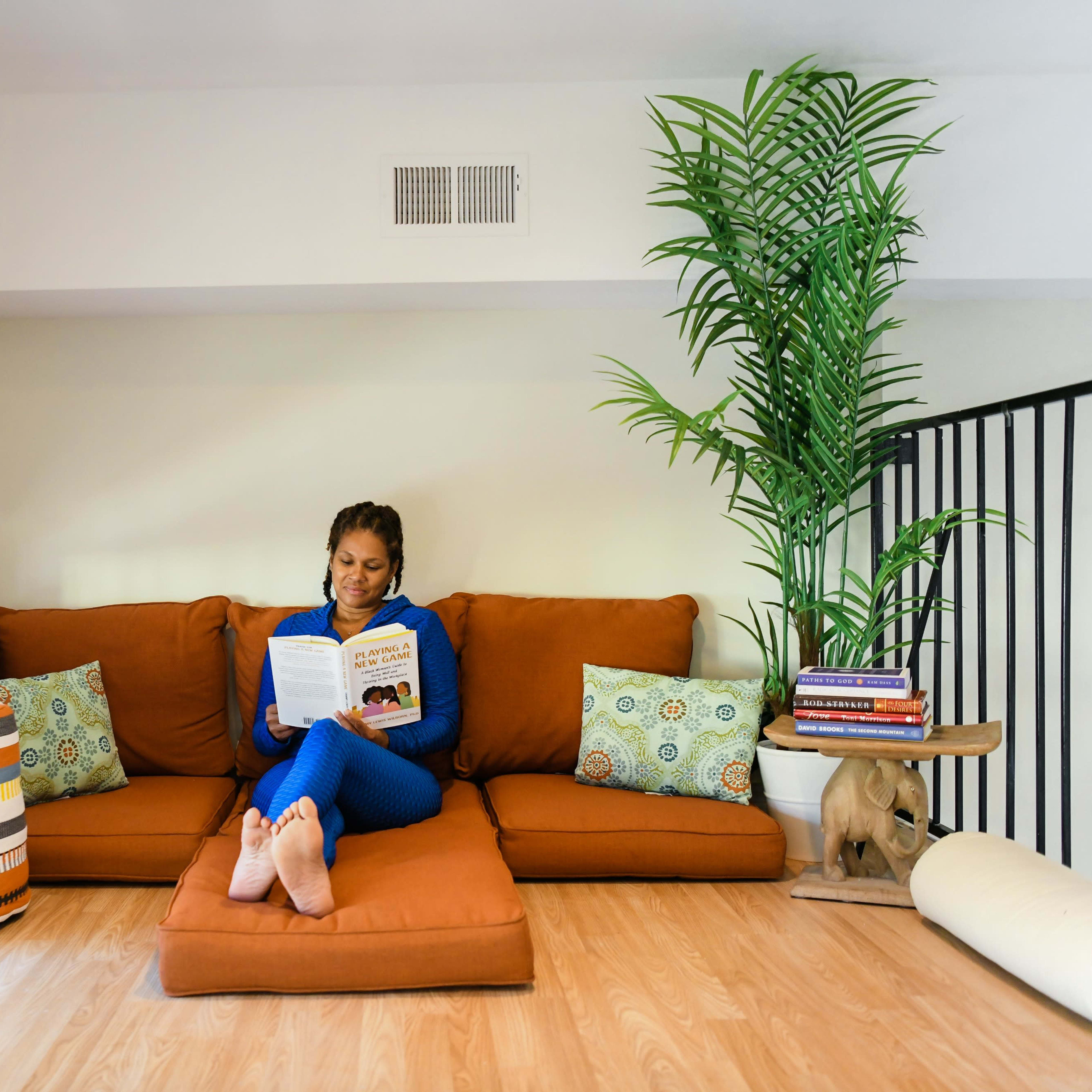 A person sits on a brown sofa, reading a book in a well-lit room with a plant and colorful decor.