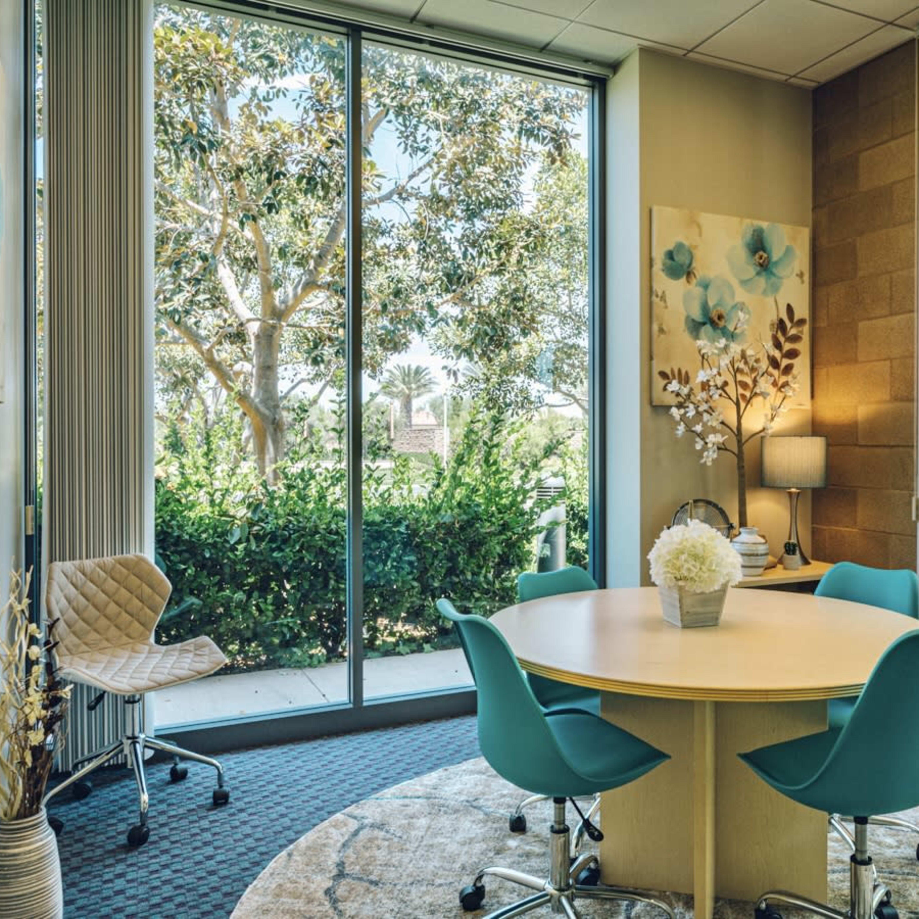 A small meeting room features a round wooden table surrounded by teal chairs, with large windows offering a view of greenery outside.