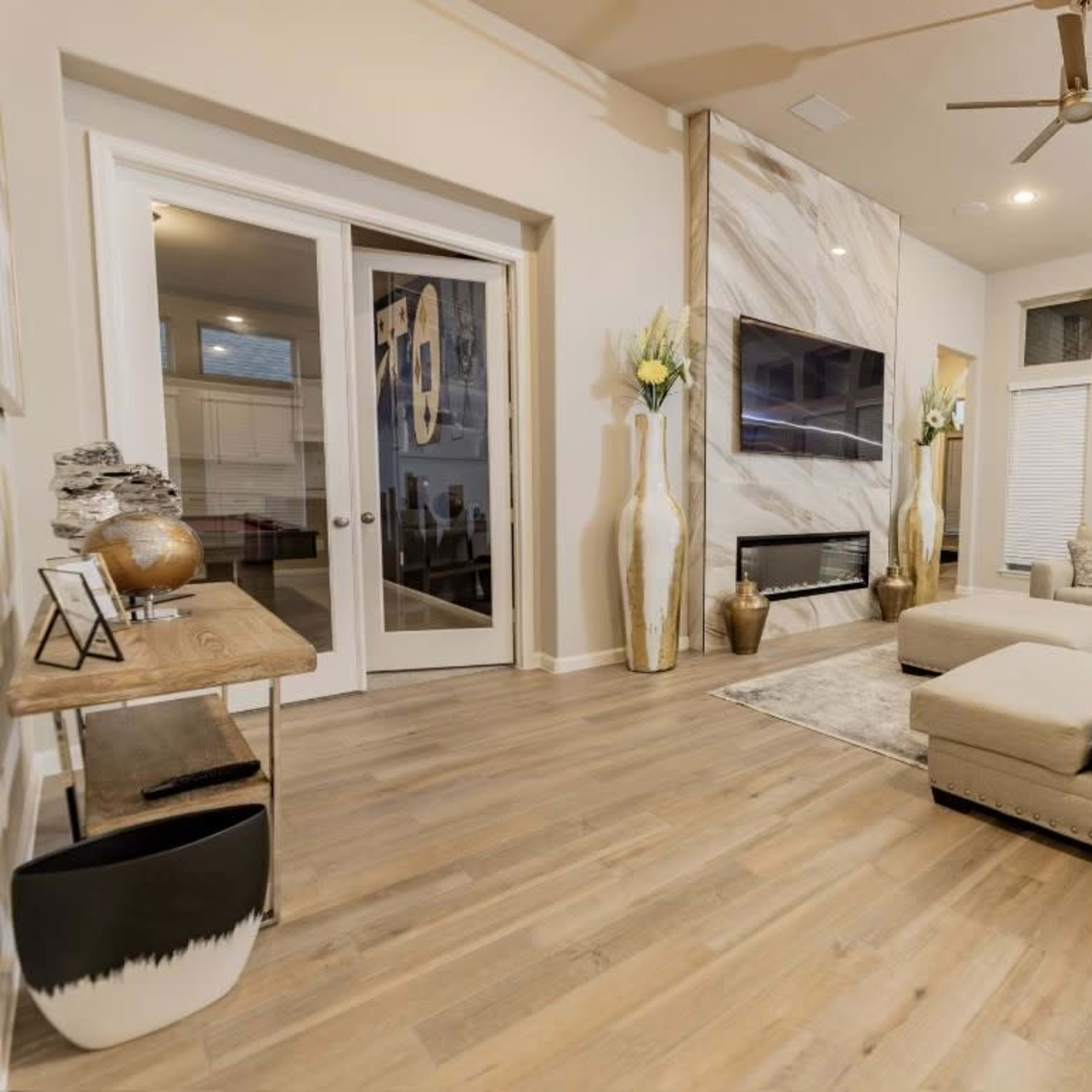 The living room features a beige sectional sofa, a wooden coffee table, and a wall-mounted television above a modern fireplace surrounded by decorative vases and large windows.