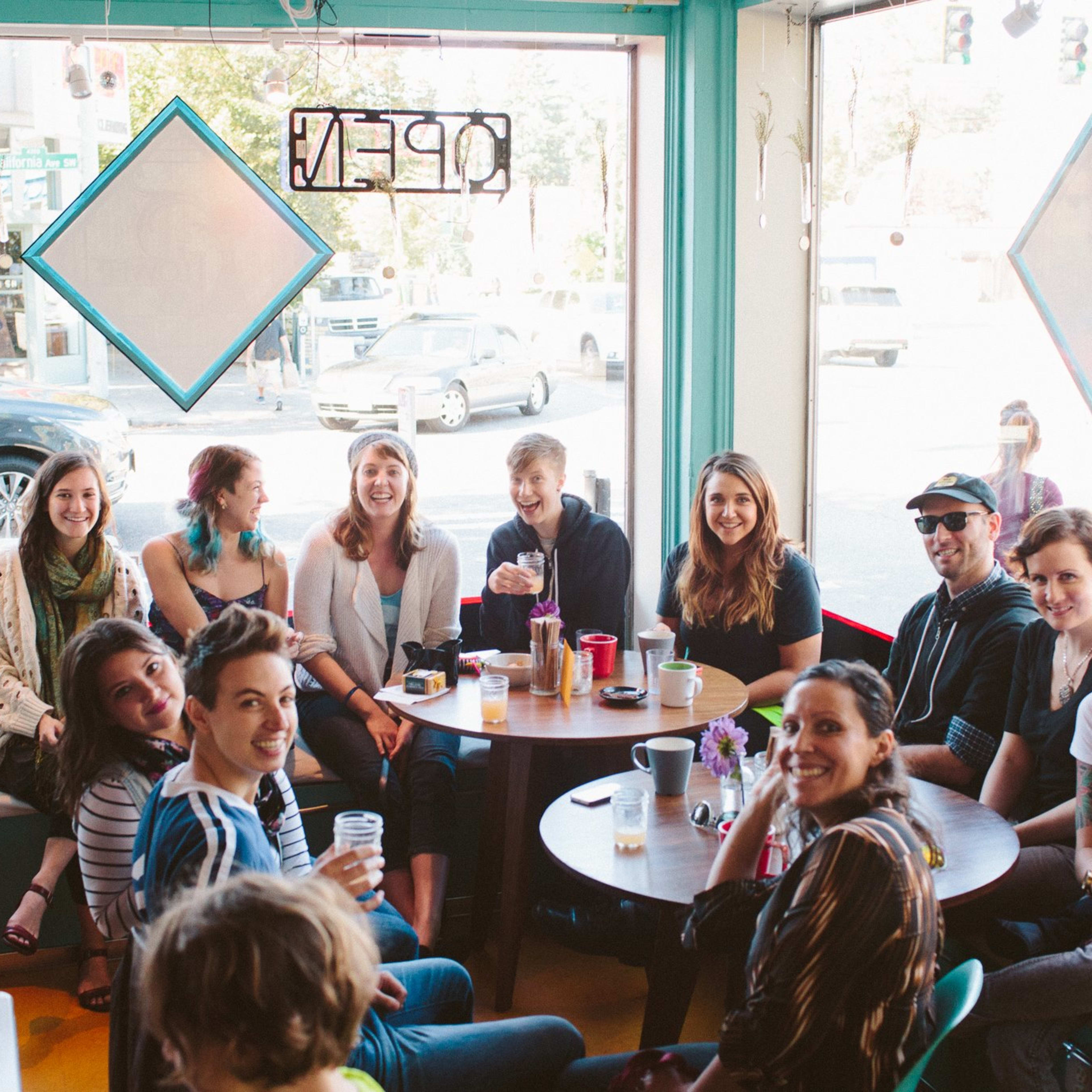 A group of people sits around a table in a brightly colored café, chatting and enjoying drinks while light filters in through large windows.