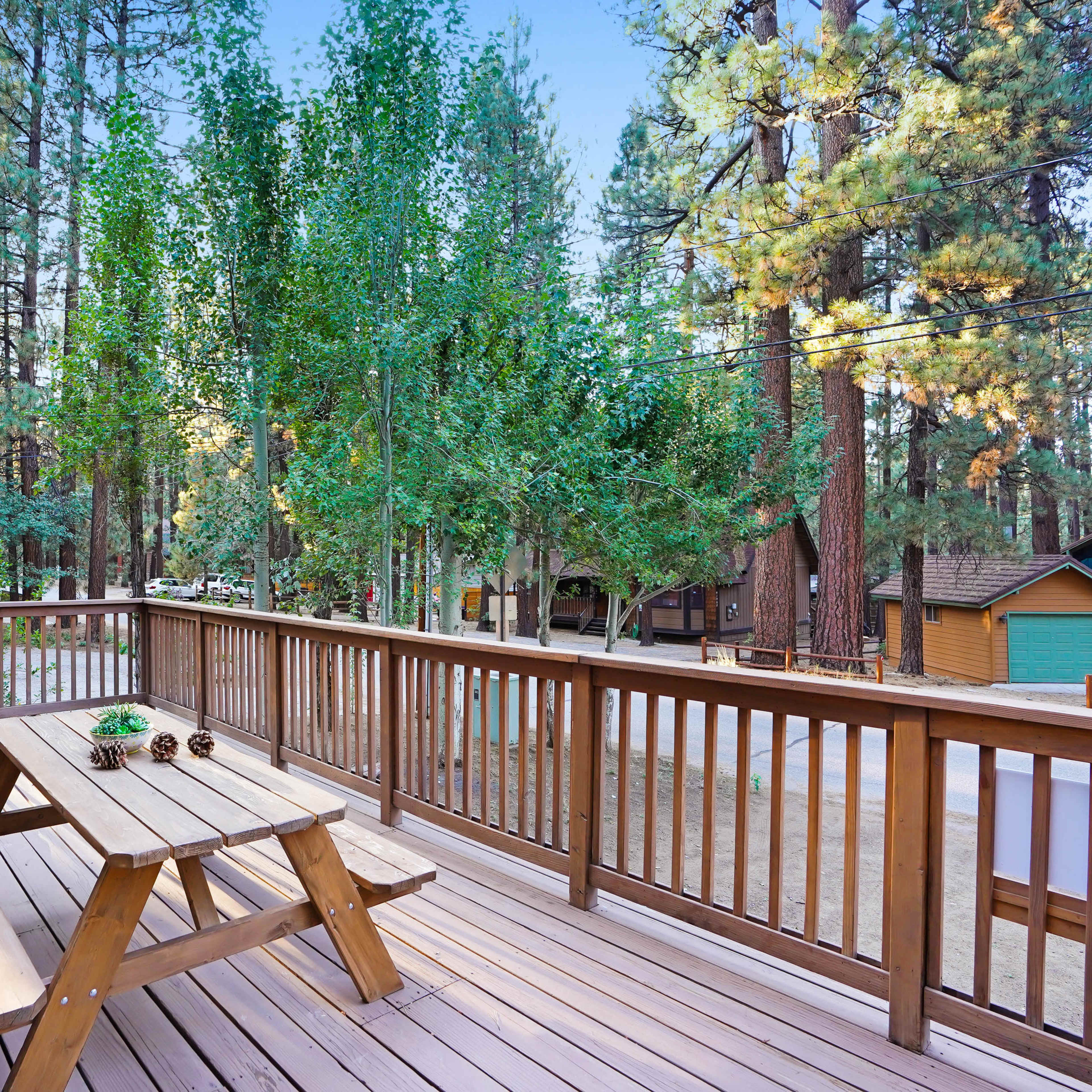 A wooden deck with a picnic table and chairs is surrounded by tall trees and cabins in a forested area.