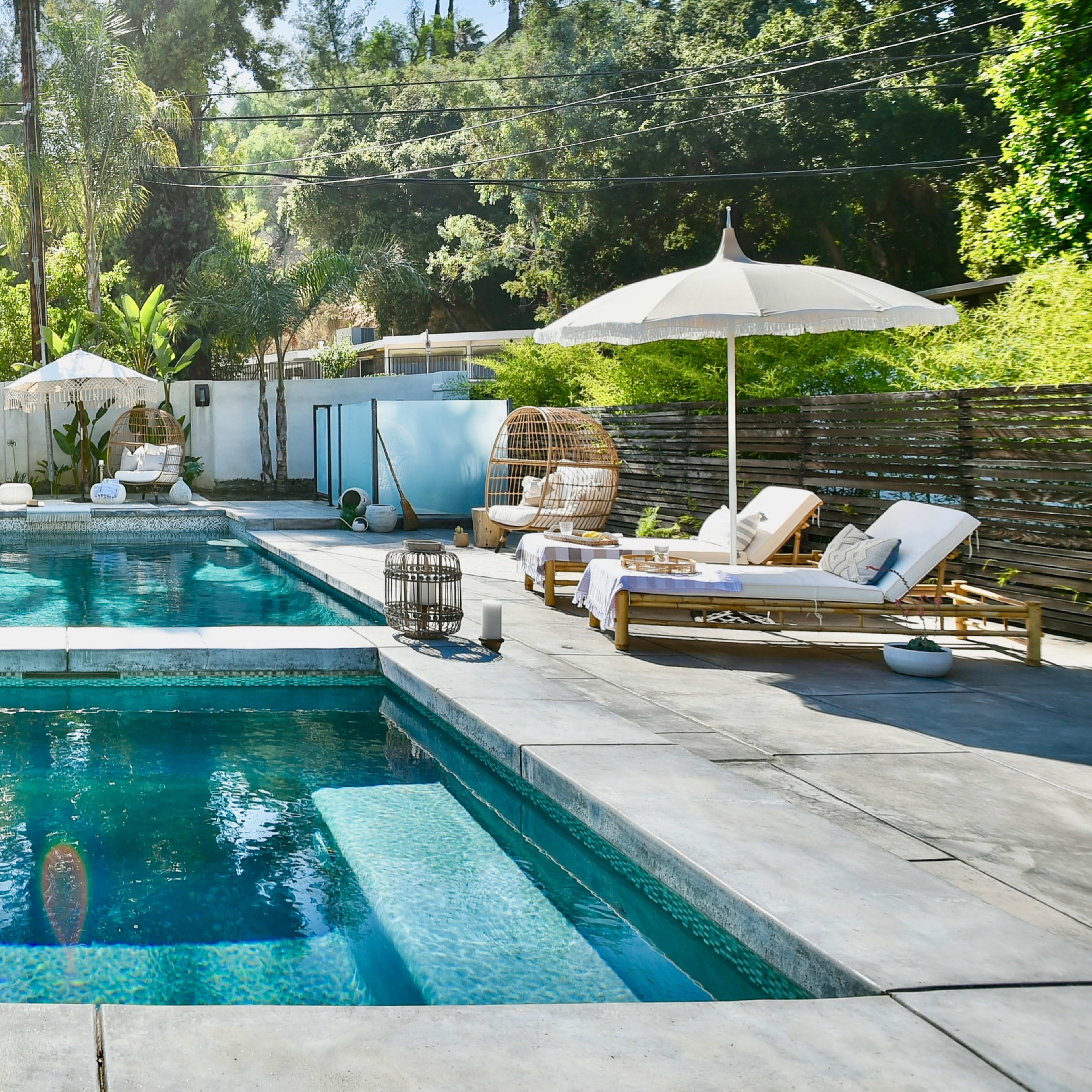 A modern backyard featuring a swimming pool with a separate spa, surrounded by tropical plants, lounge chairs, and an umbrella.