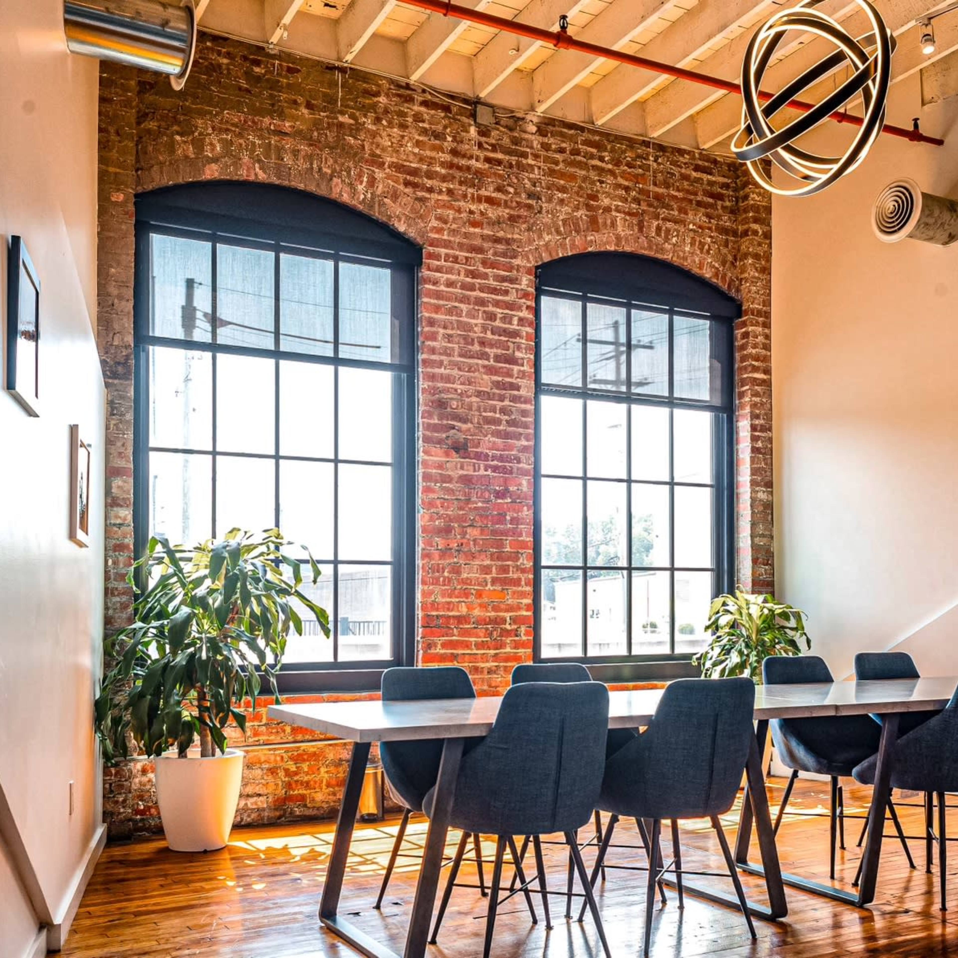 A brightly lit meeting room features a rectangular table surrounded by dark chairs, with large windows and exposed brick walls creating a modern aesthetic.