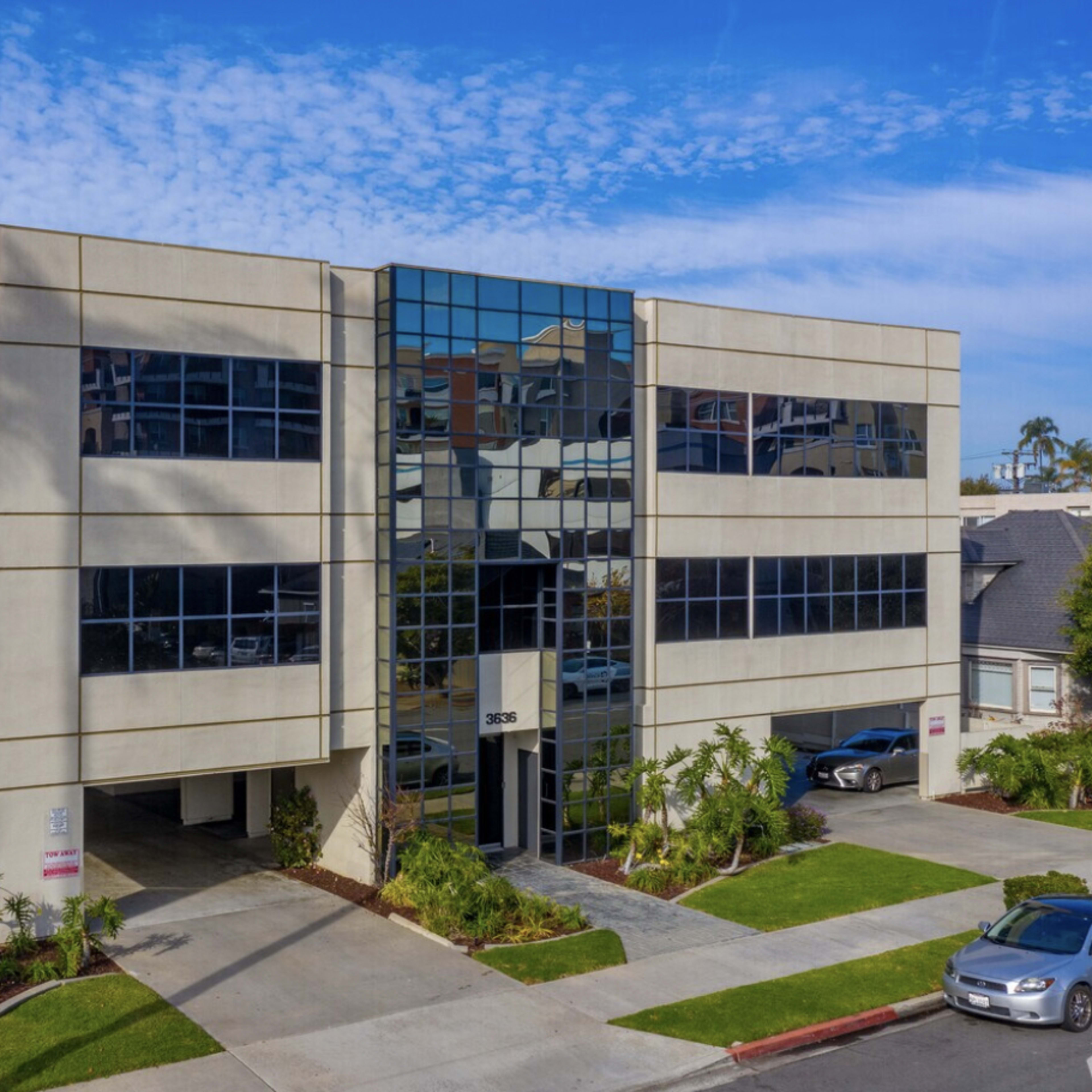 A modern, rectangular building with large glass windows and a landscaped entrance is situated on a street lined with palm trees.