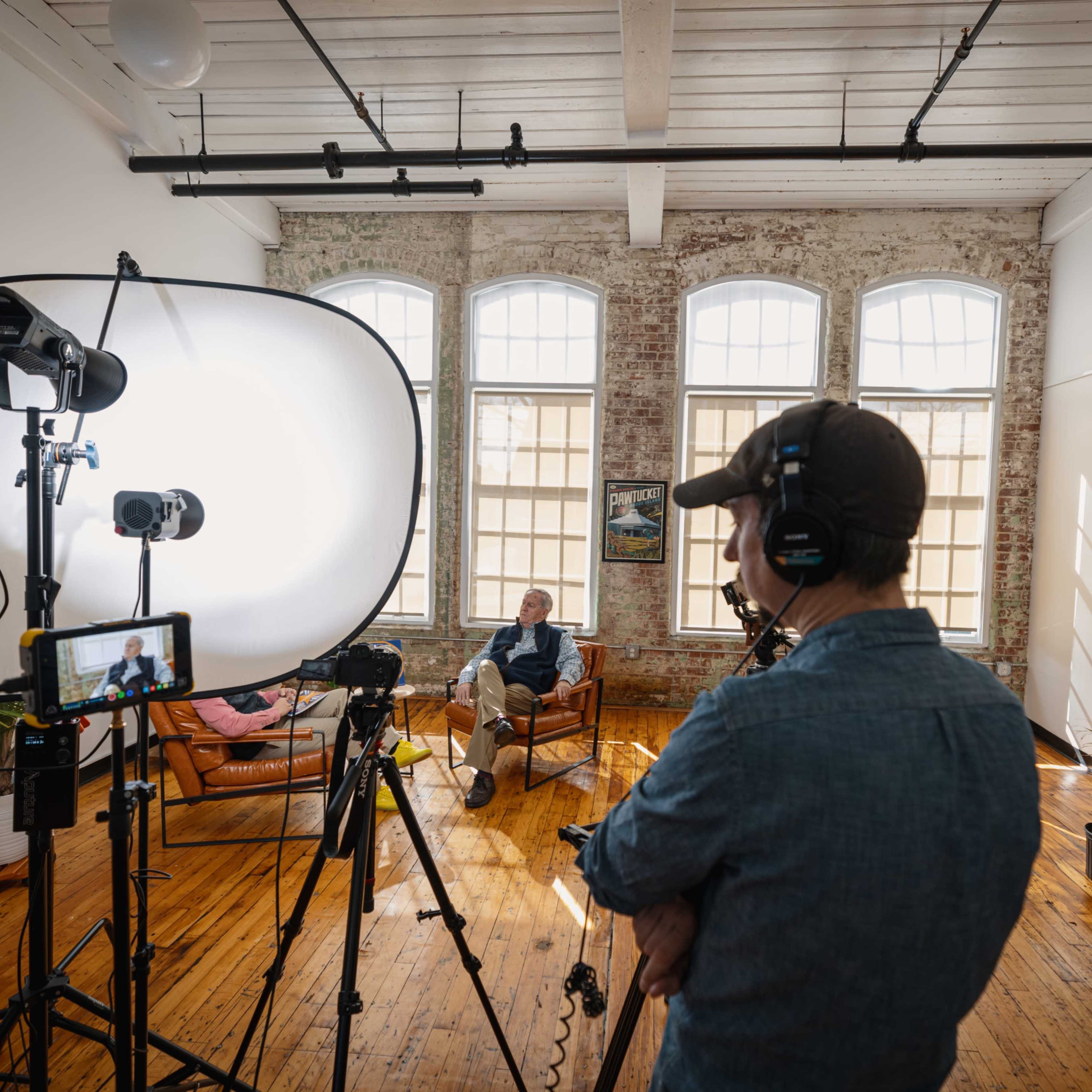 A person wearing a headset stands in front of a camera setup, while another person sits on a couch in a well-lit, modern room with large windows and wooden floors.