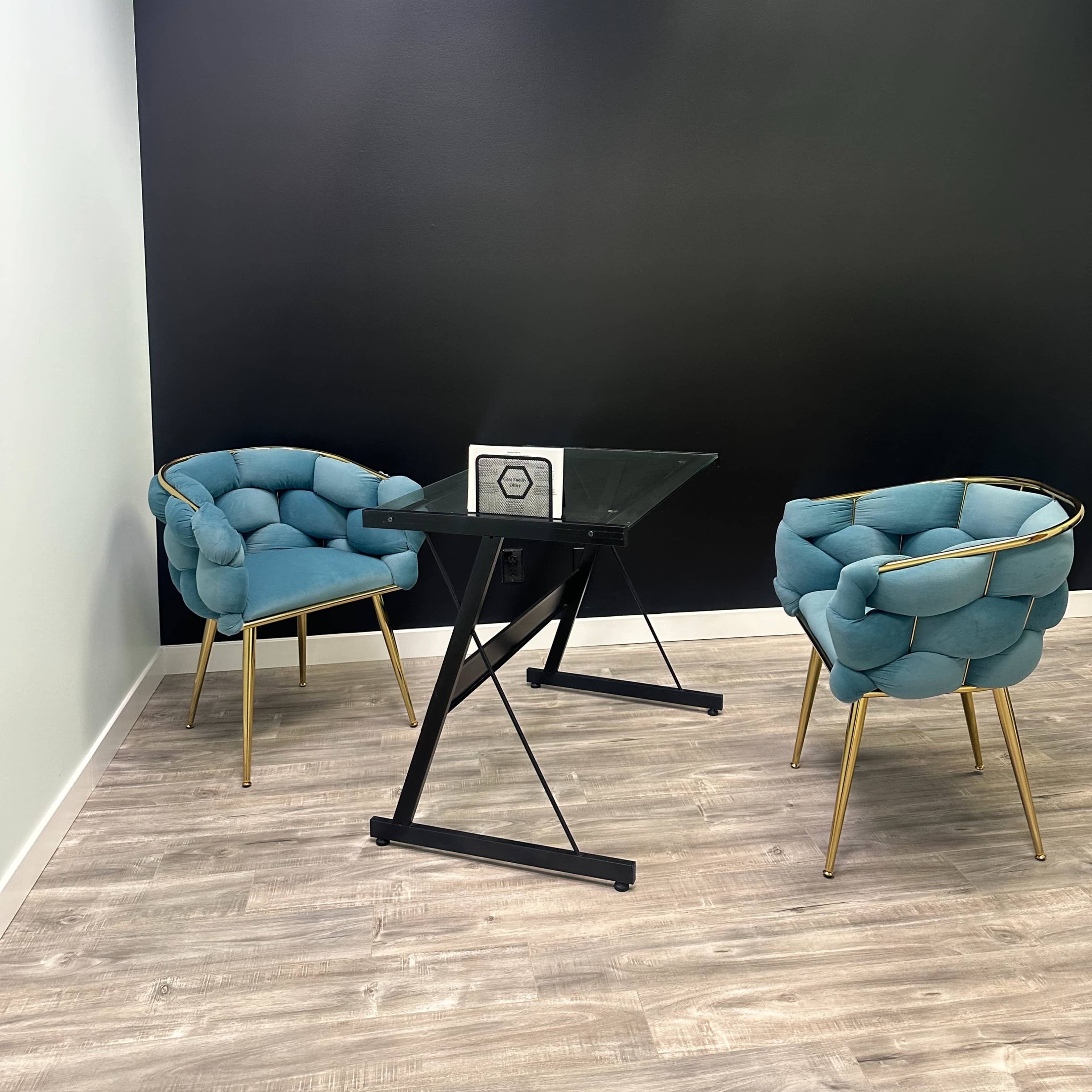 A modern office space features two teal chairs and a glass-top table against a black wall.