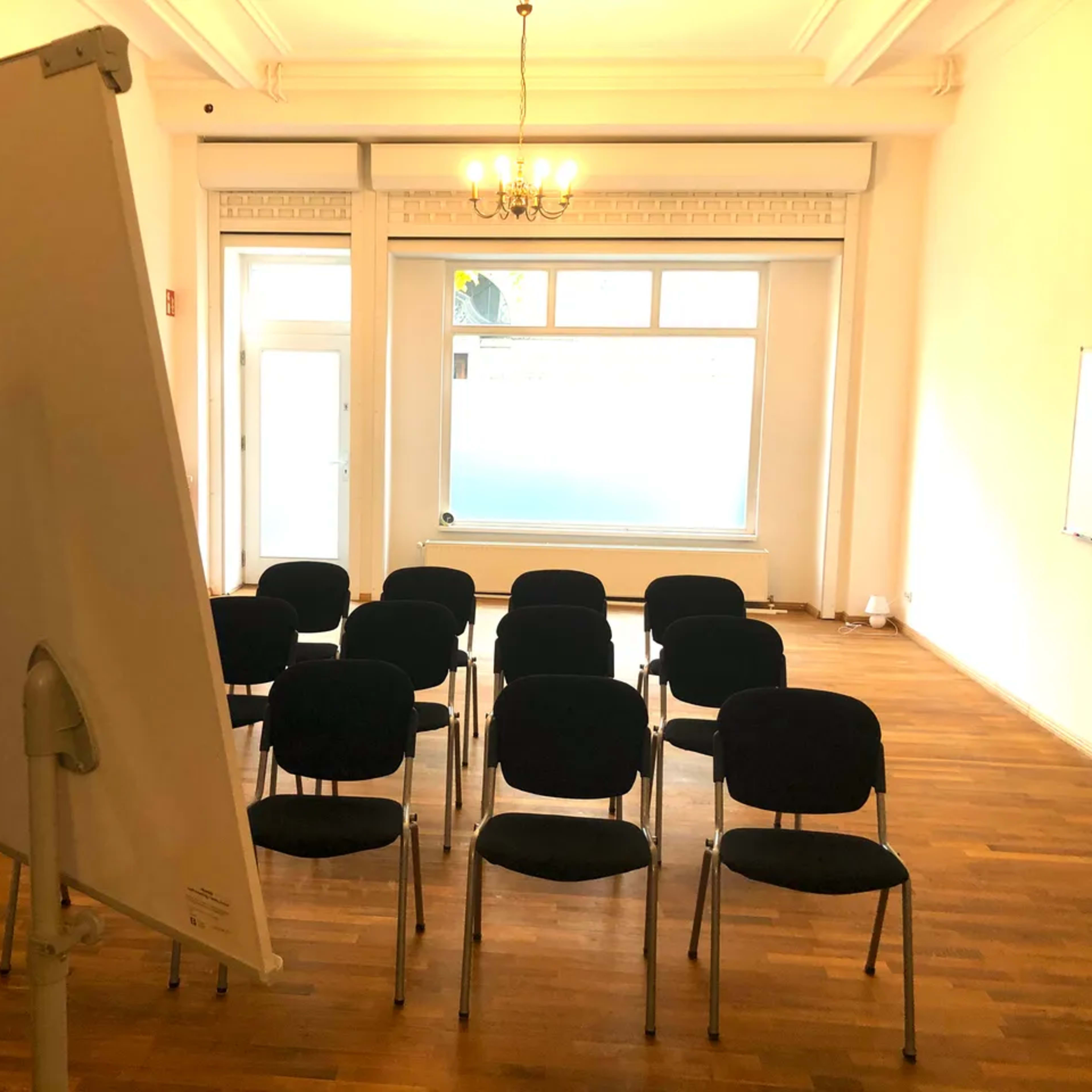 A spacious room with wooden flooring contains several chairs arranged in rows facing a flip chart and a large window.