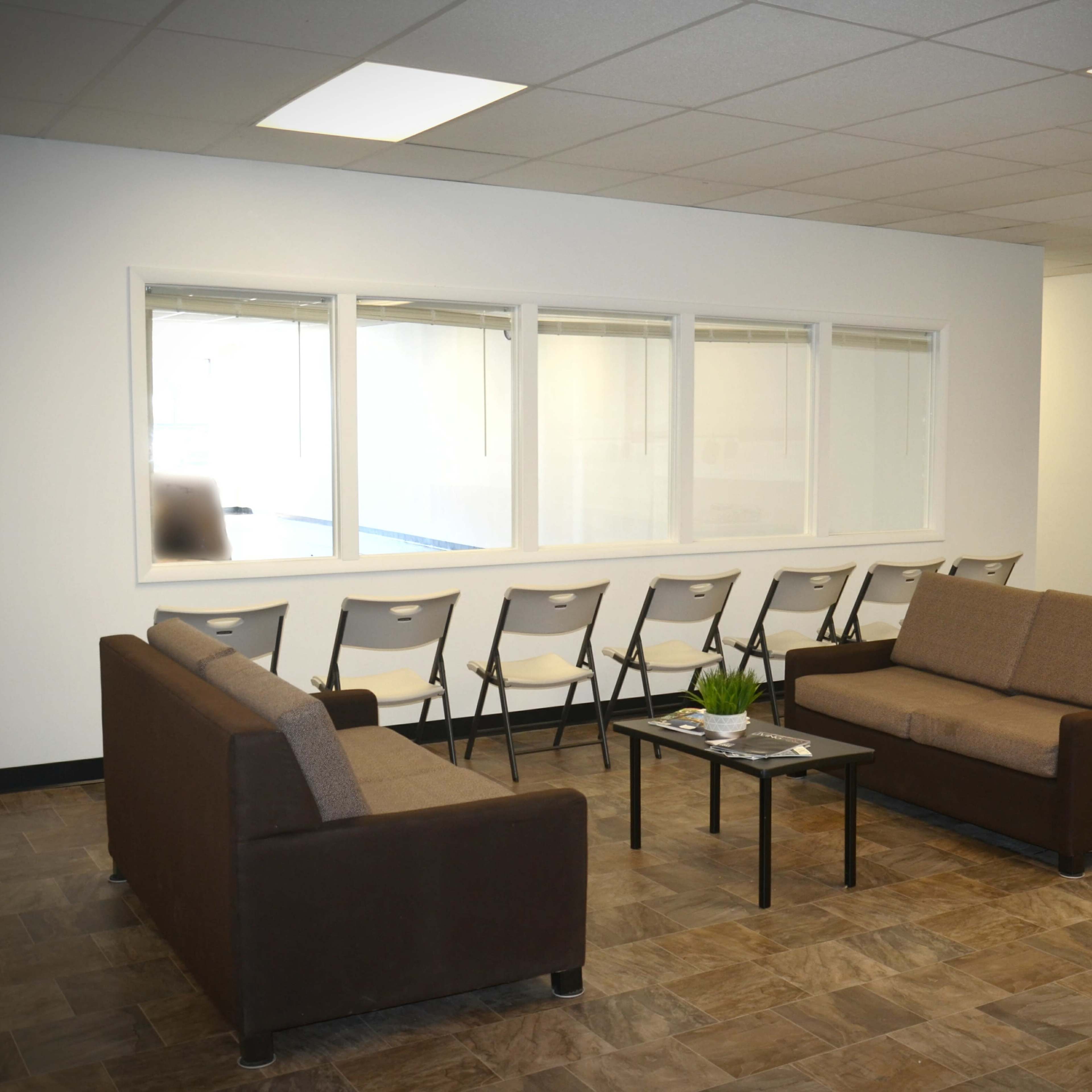 A waiting area with two brown sofas, a small table, and a row of folding chairs positioned against a wall with large windows.