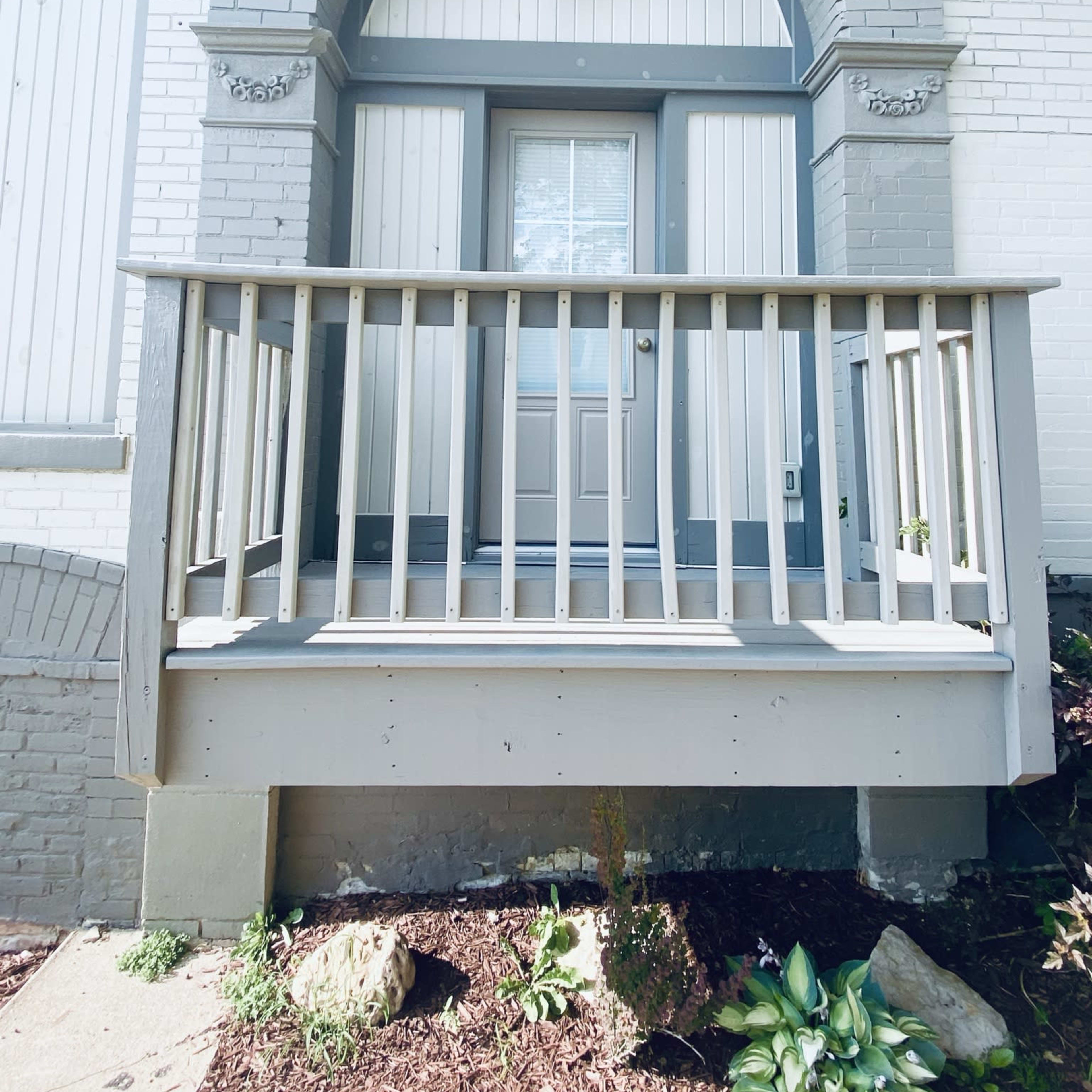 A small front porch with a wooden railing leading up to a door framed by an arched structure on a brick building.