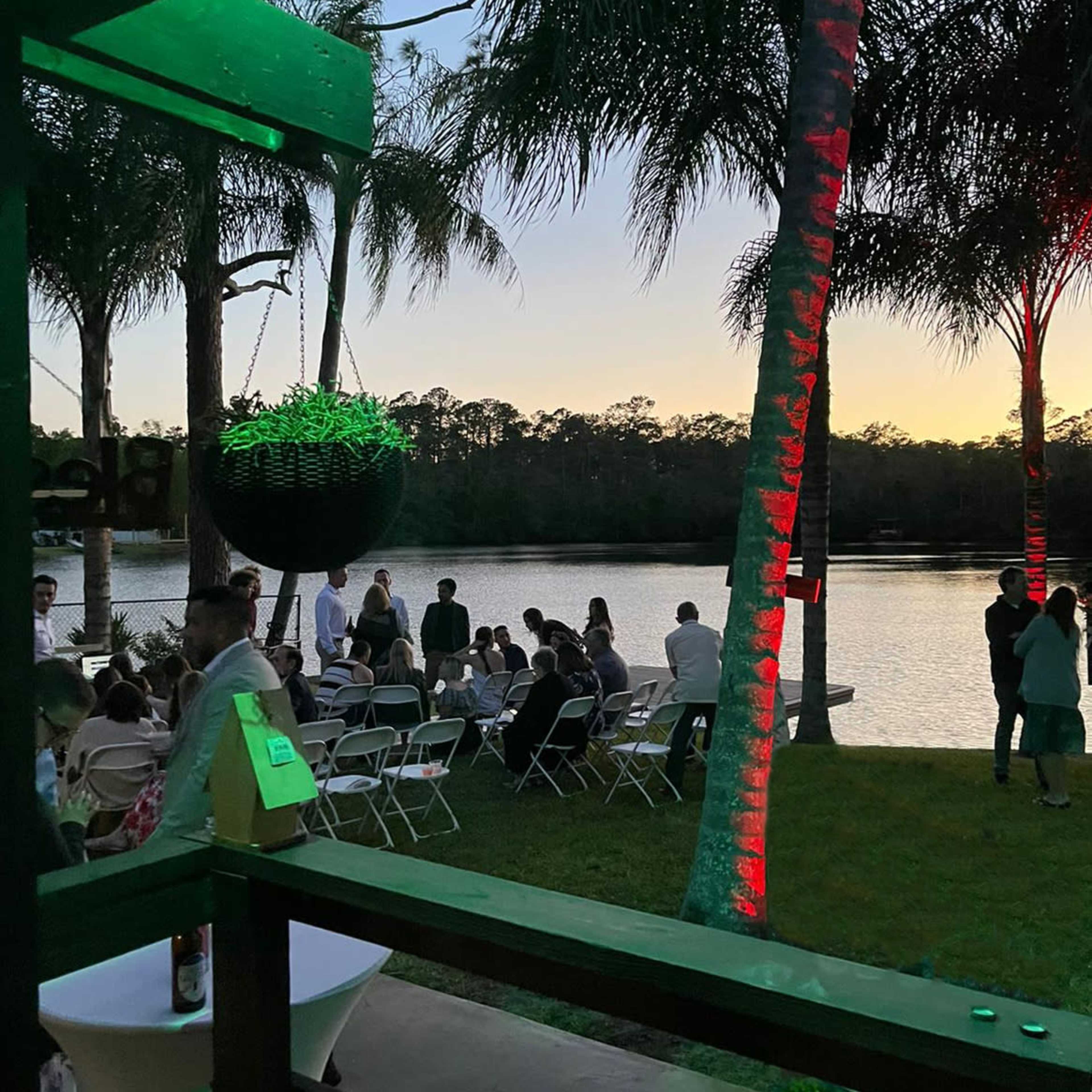 A group of people gathers along the shore of a lake at sunset, with palm trees and chairs arranged for an event.