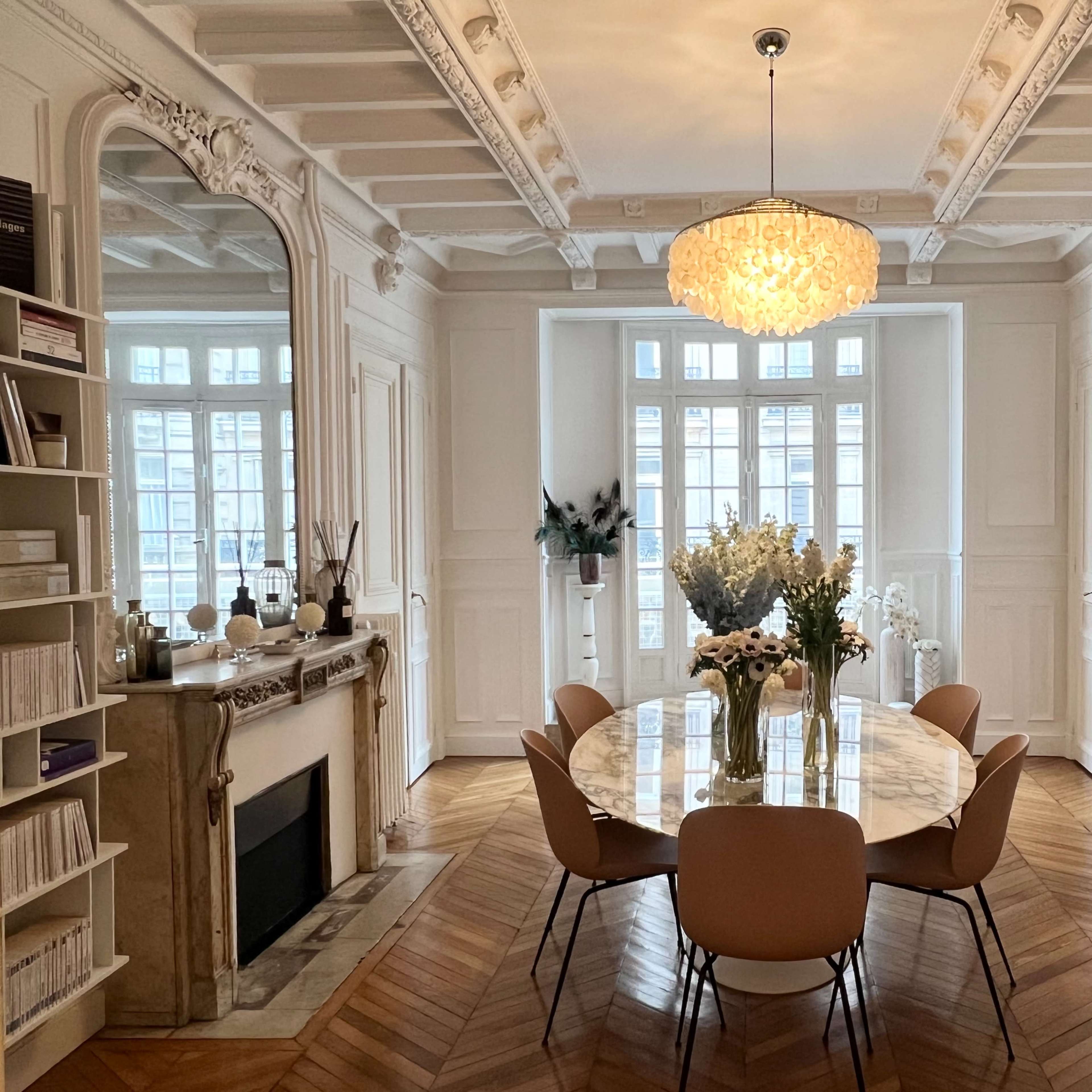 A modern dining room features a round marble table surrounded by beige chairs, with a large chandelier overhead and decorative elements on the walls.