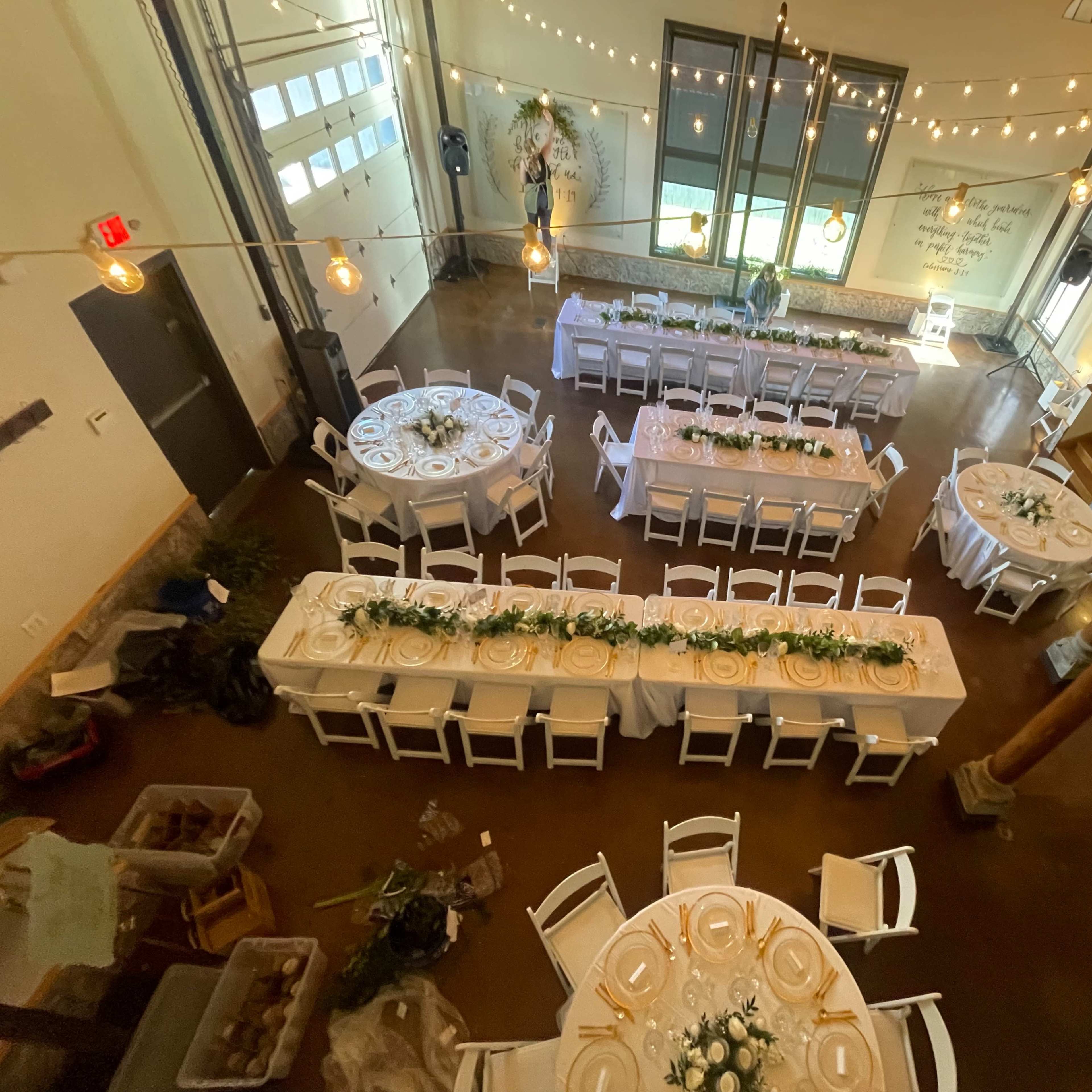 A spacious event hall arranged with multiple circular and rectangular tables decorated with white tablecloths and floral centerpieces, illuminated by string lights.