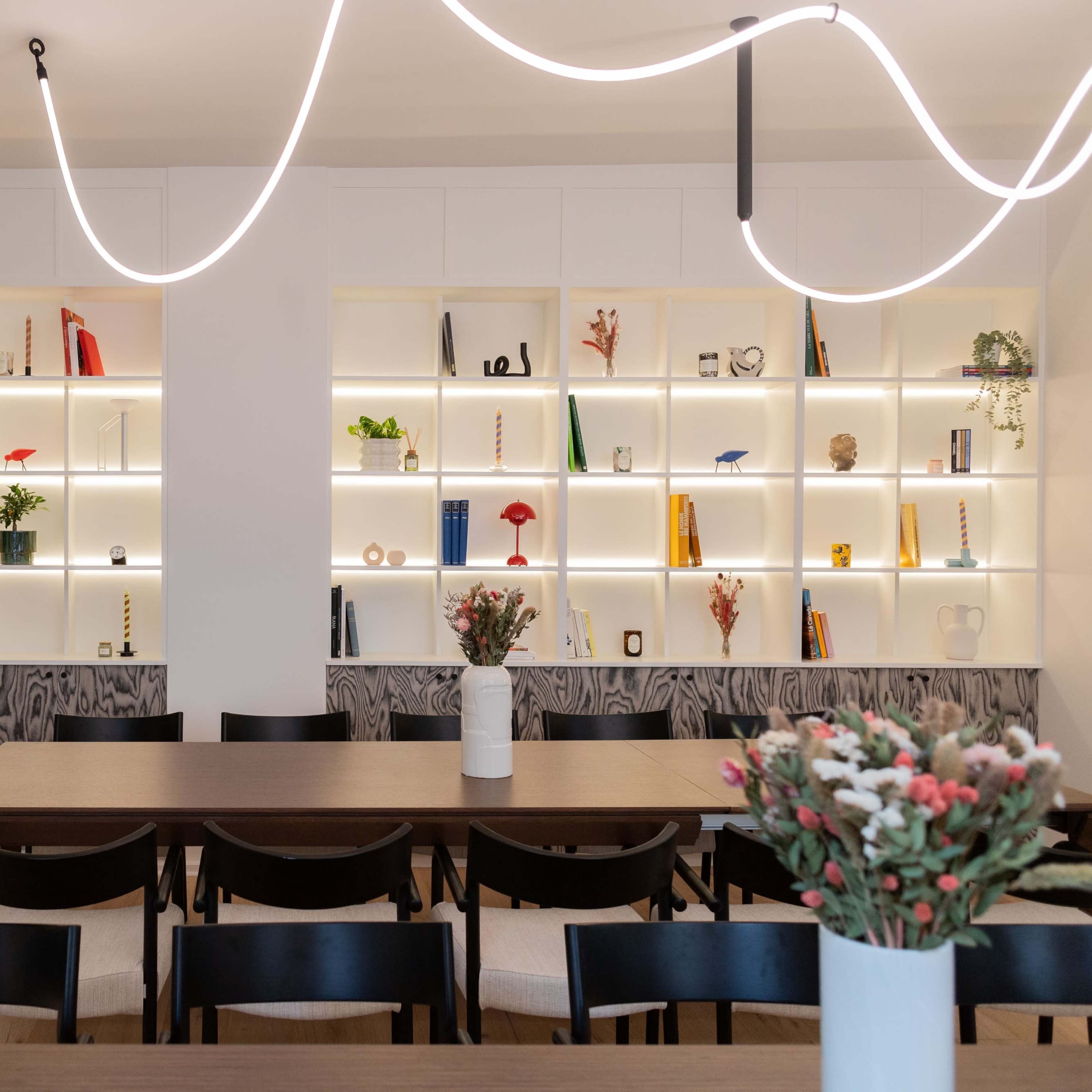 A modern meeting room features a long table surrounded by black chairs, with a well-lit bookshelf displaying decorative items and plants in the background.
