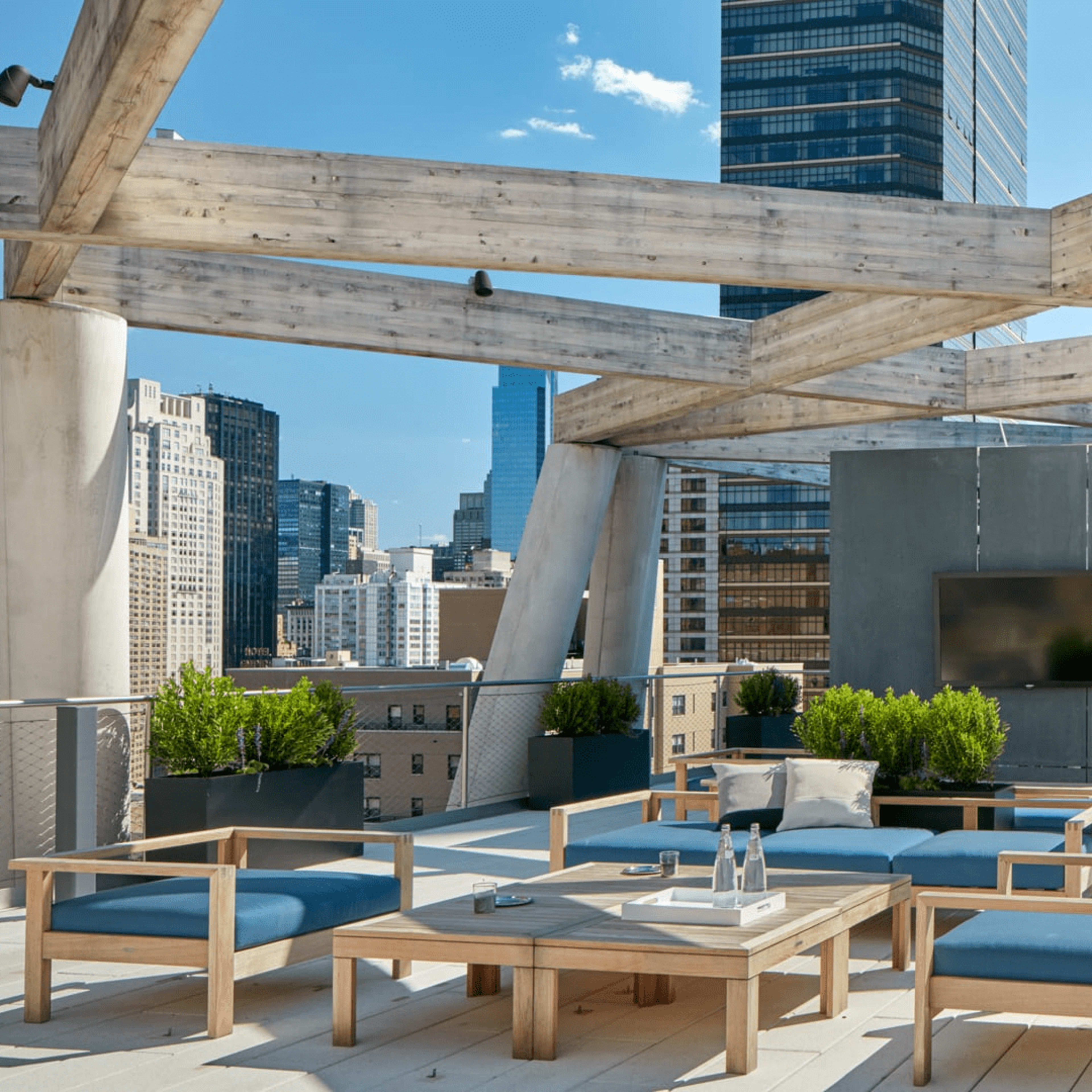 The rooftop terrace features wooden beams, modern seating, and planters, overlooking a city skyline with tall buildings.