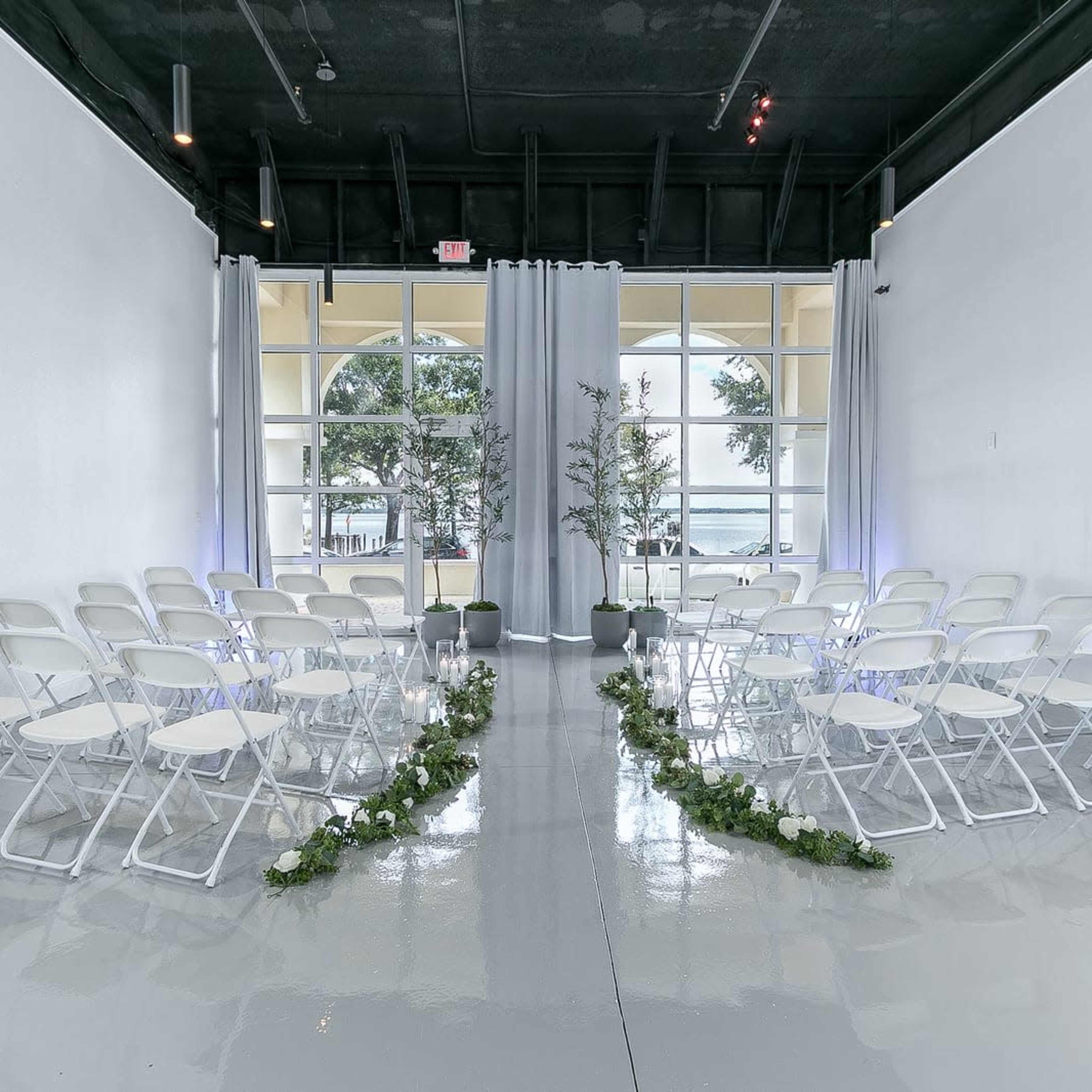 A wedding ceremony setup with white chairs arranged in two rows facing a draped area, surrounded by greenery and floral decorations.