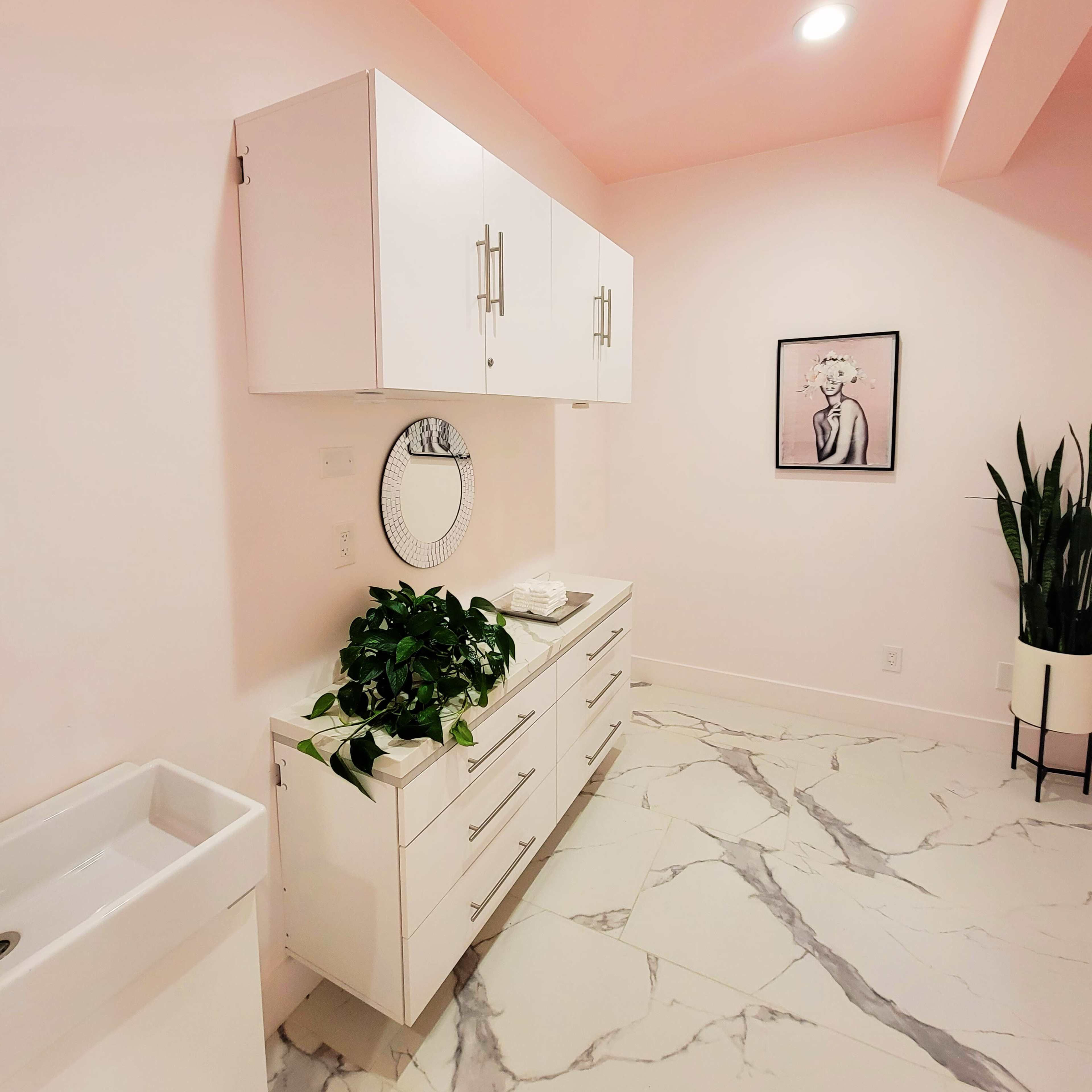 A modern bathroom features a white vanity with cabinets, a round mirror, a plant, and marble flooring against a pink wall.
