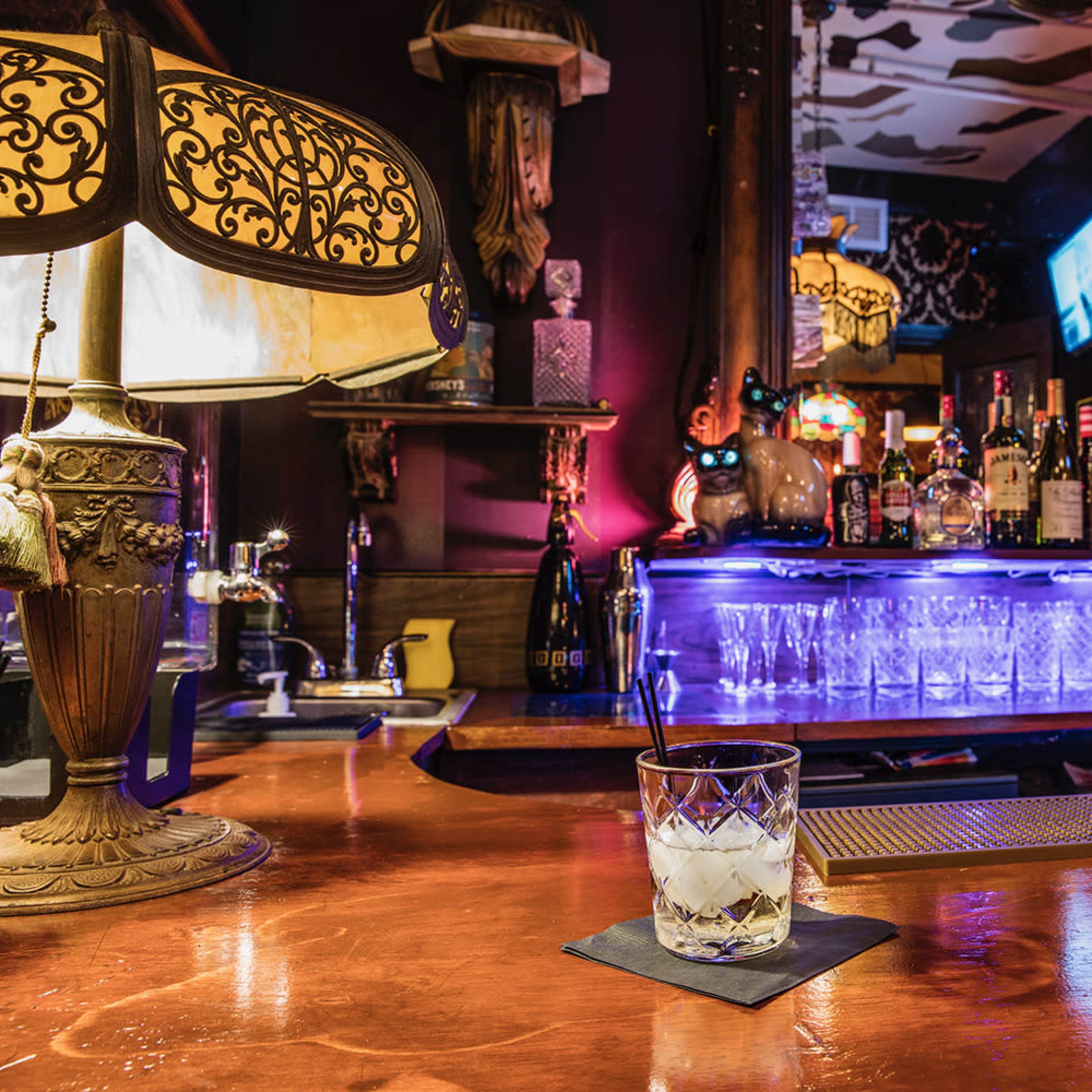A well-lit bar scene features a glass of cocktail on the counter in front of a backlit shelf stocked with various liquor bottles.