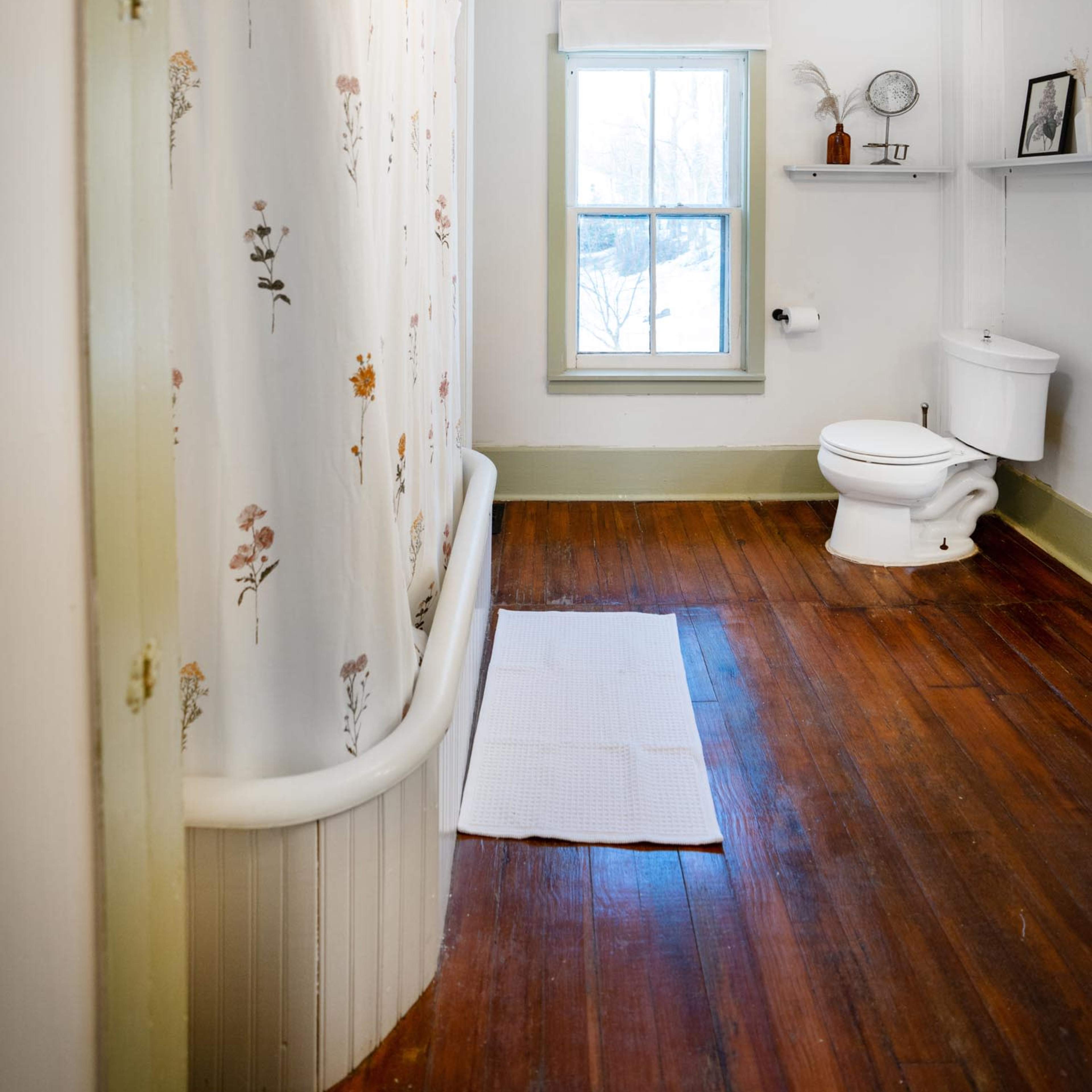 A bathroom featuring a clawfoot tub with a flower-patterned shower curtain, a white toilet, and wooden flooring.