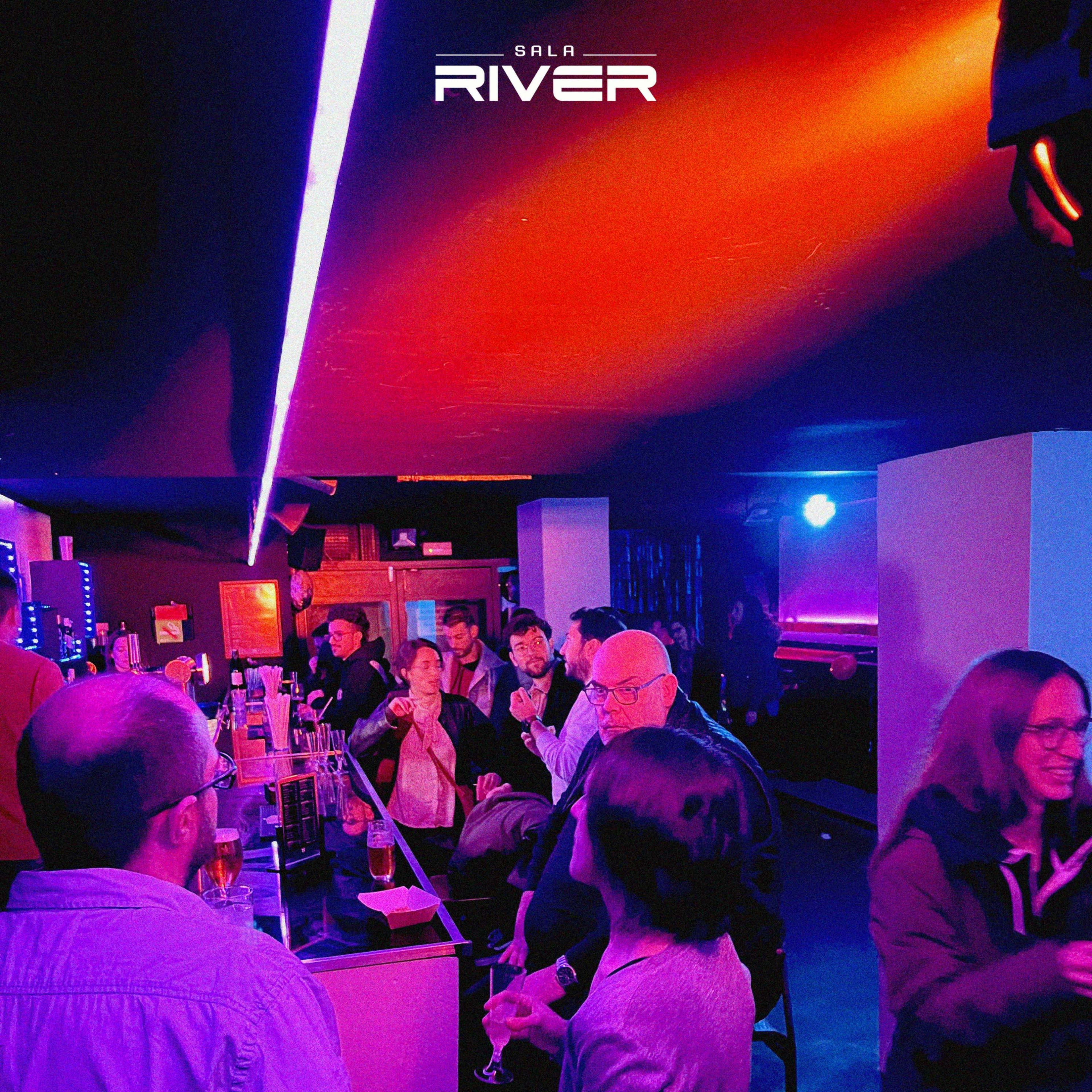 A lively bar scene with patrons socializing and drinks on the counter, illuminated by colorful lights.