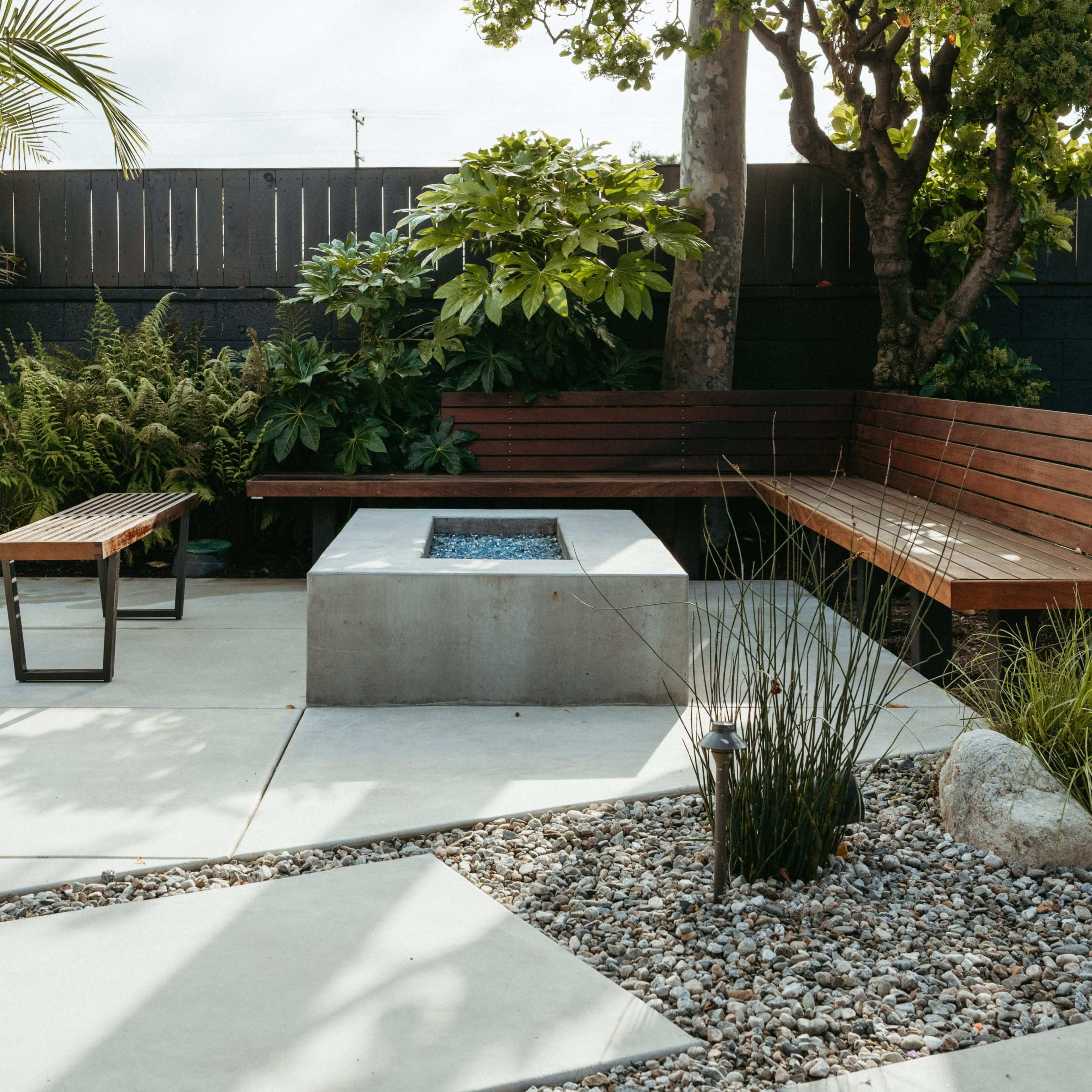 A modern outdoor seating area featuring a concrete fire pit surrounded by wooden benches, stone landscaping, and greenery.