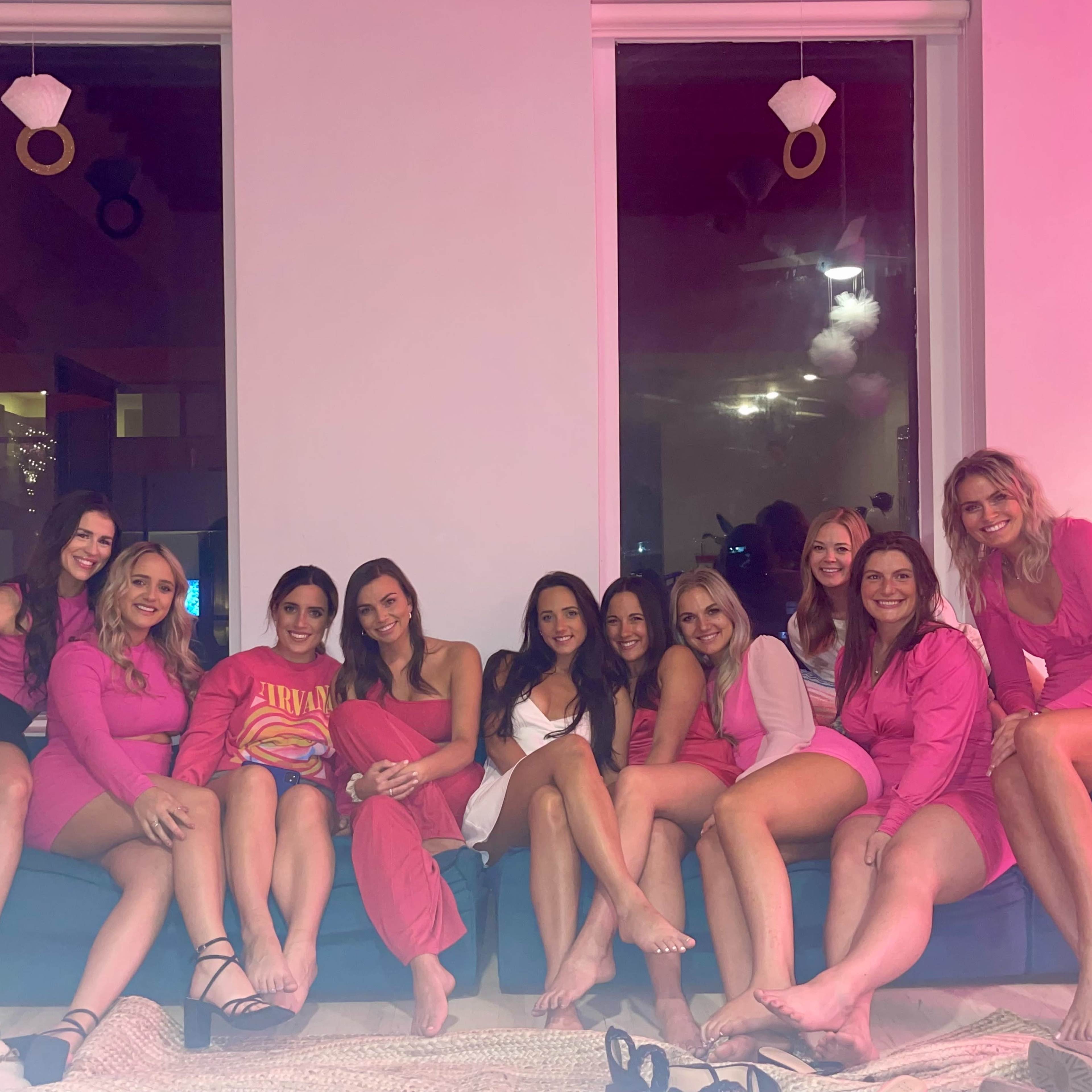 A group of ten women in matching outfits sits on a couch in a well-lit room with large windows and decorative elements.