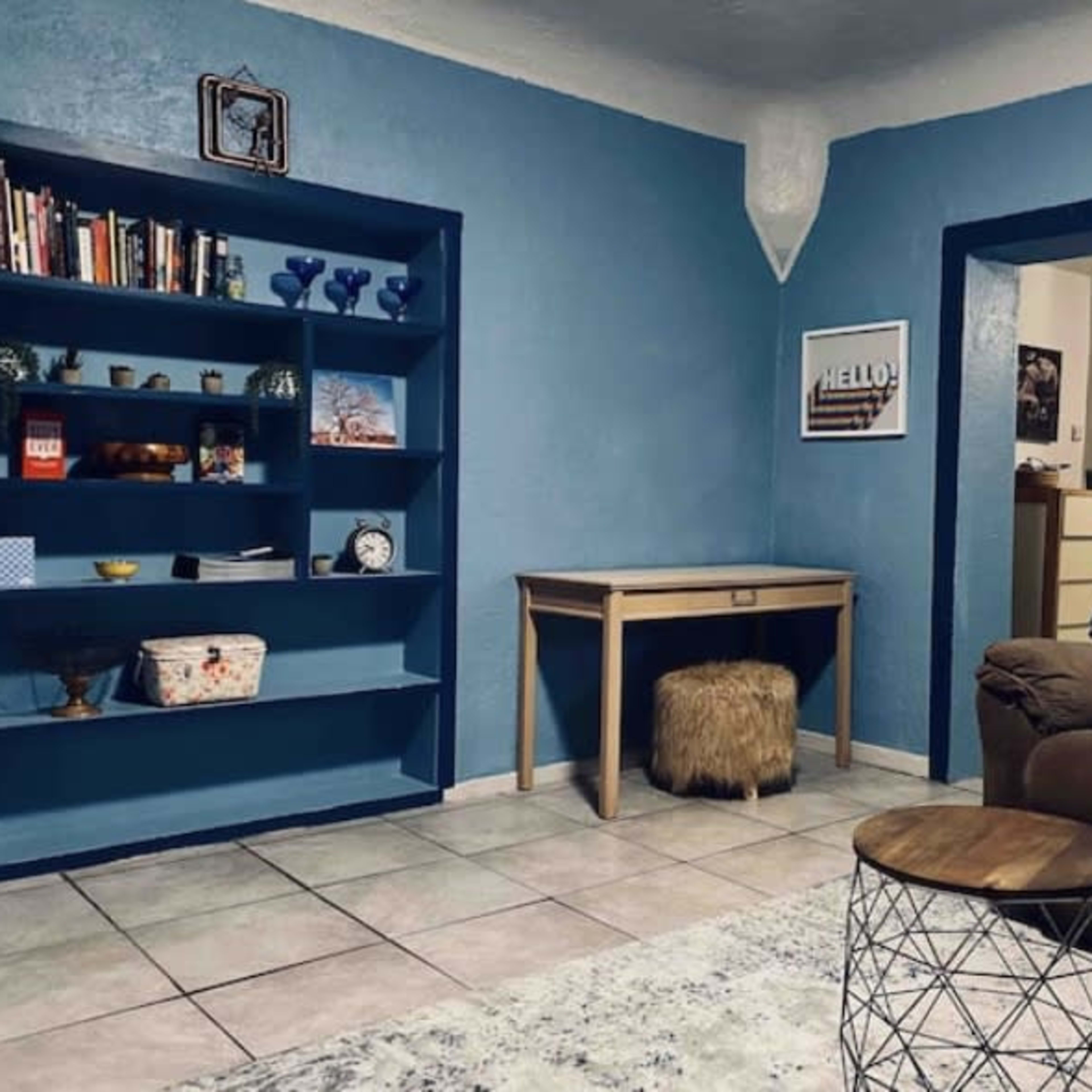 A living room with blue walls, a bookshelf filled with books and decorative items, a desk, and a brown couch.