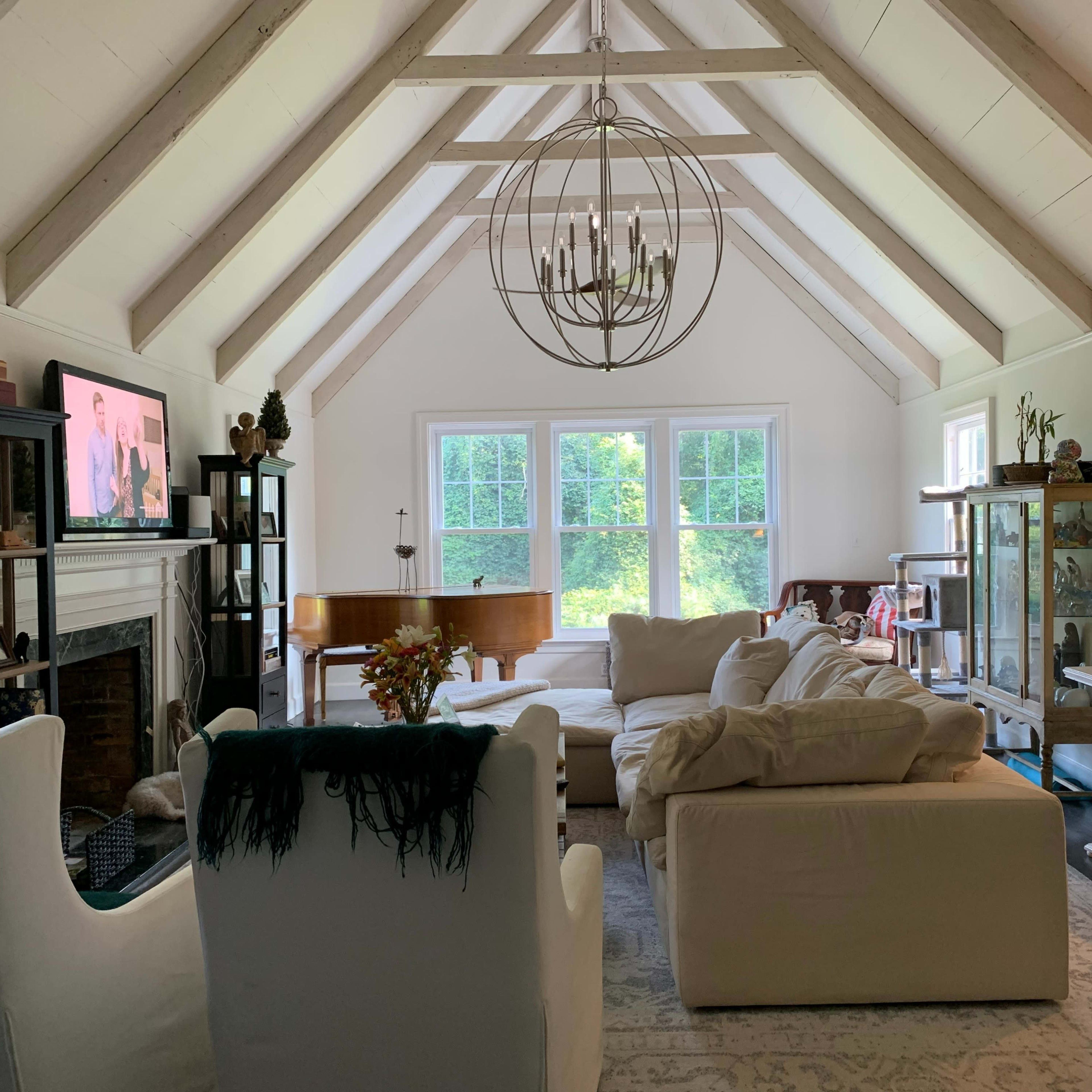 The living room features a vaulted ceiling with exposed beams, a large sectional sofa, a piano, and multiple windows allowing natural light to fill the space.