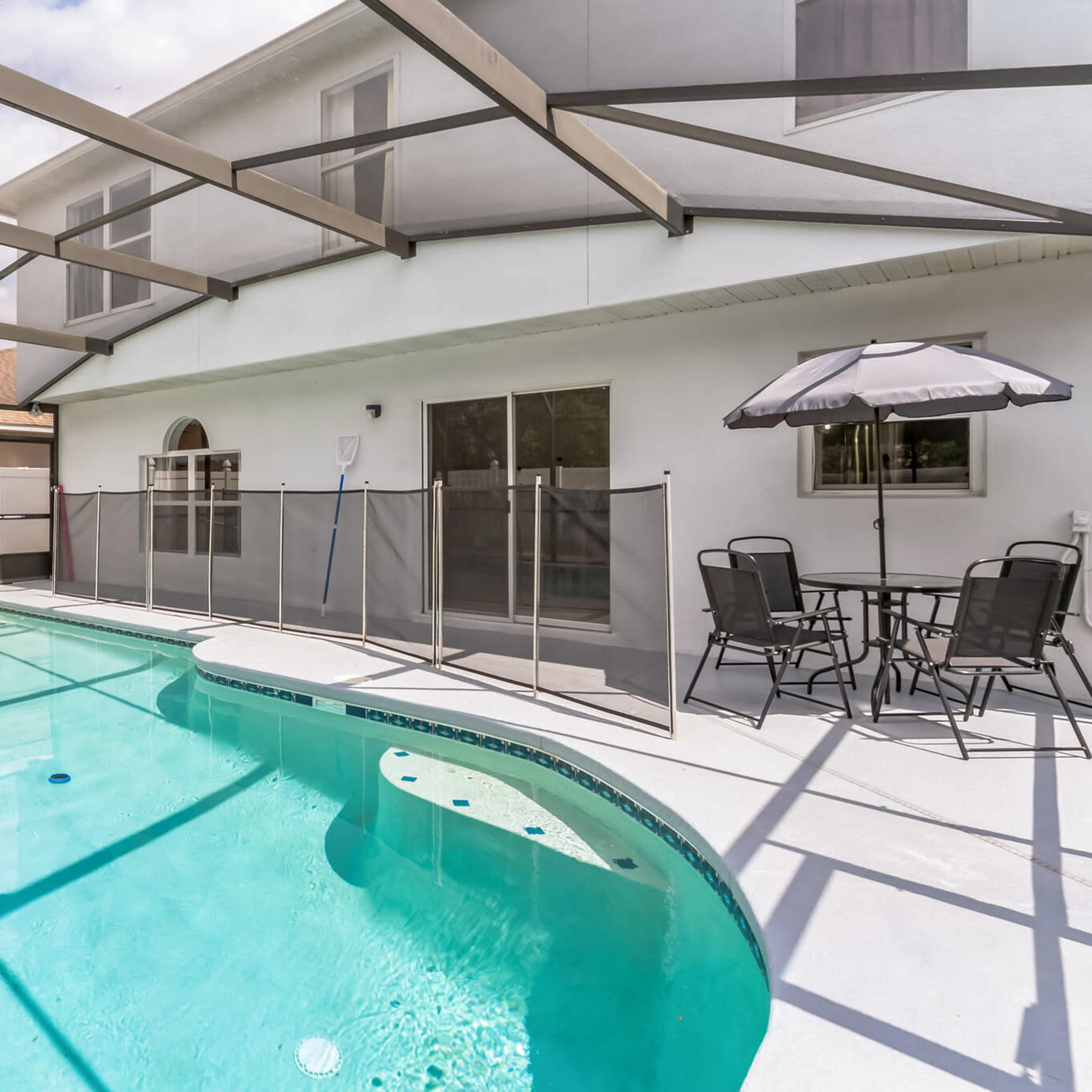 A fenced swimming pool area with a table and chairs covered by an umbrella, set against a white house and clear sky.