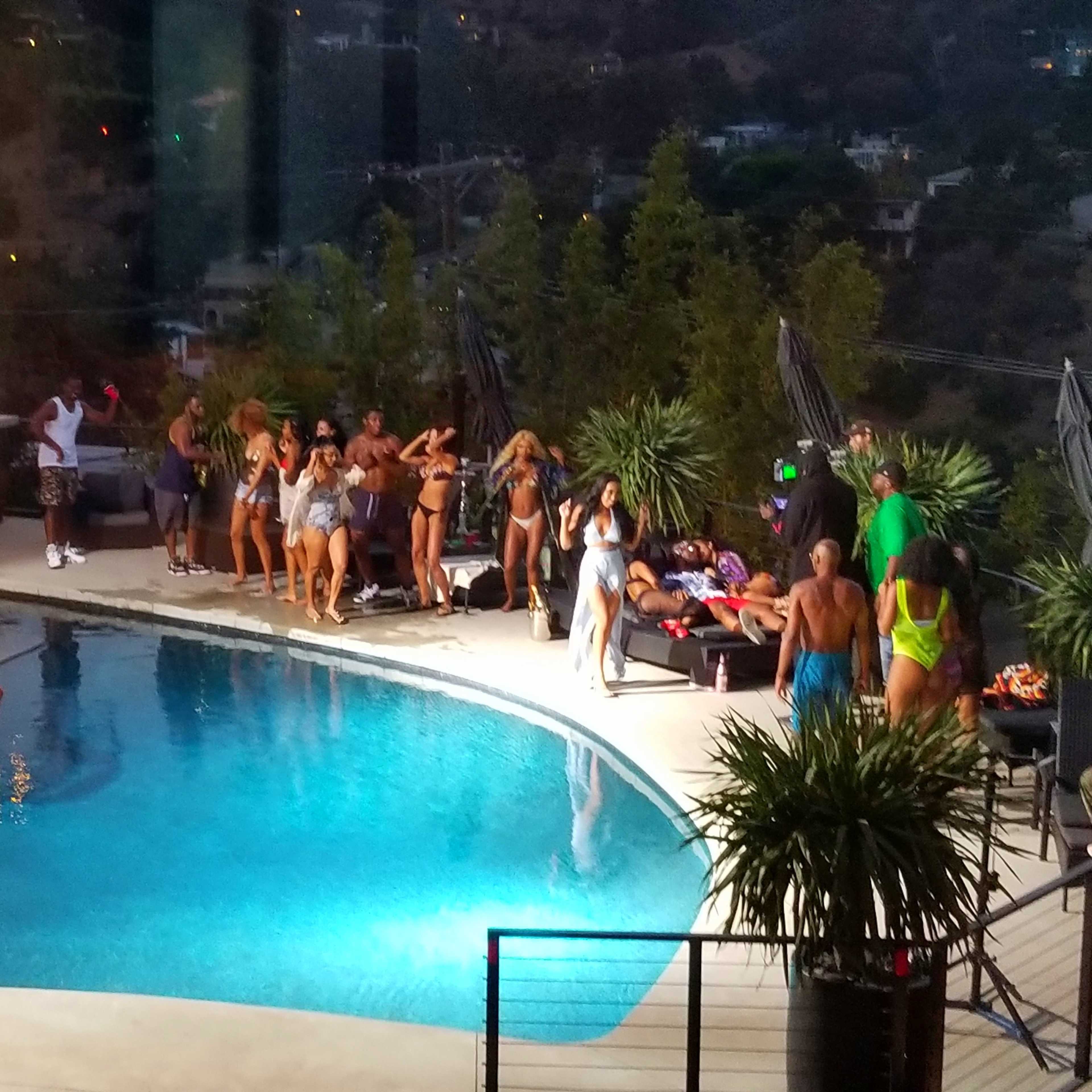 A large group of people is gathered around a poolside area featuring a fire pit, inflatable pool toys, and a backdrop of hills.