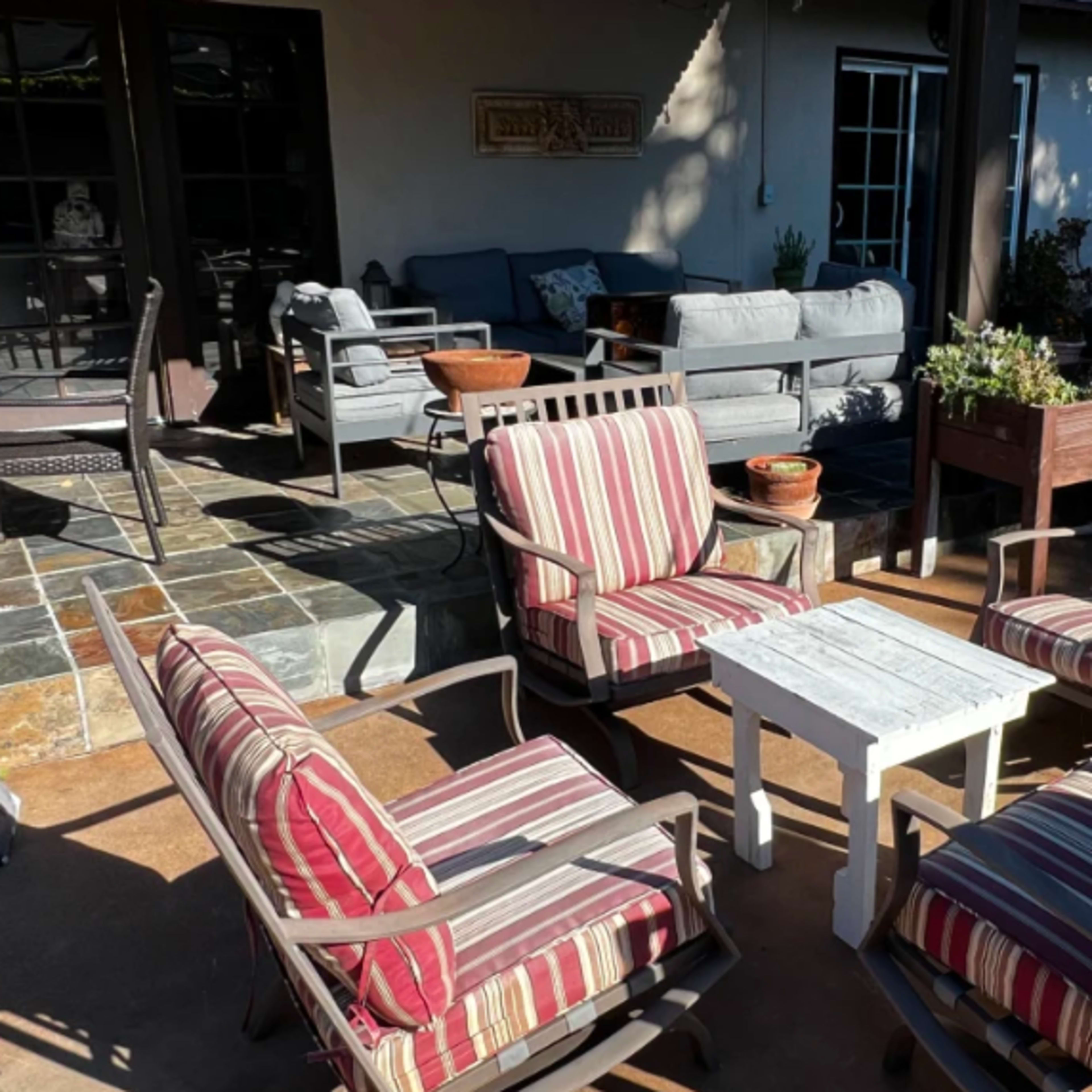 A patio area features striped armchairs arranged around a small white table, with potted plants and a shaded seating area in the background.