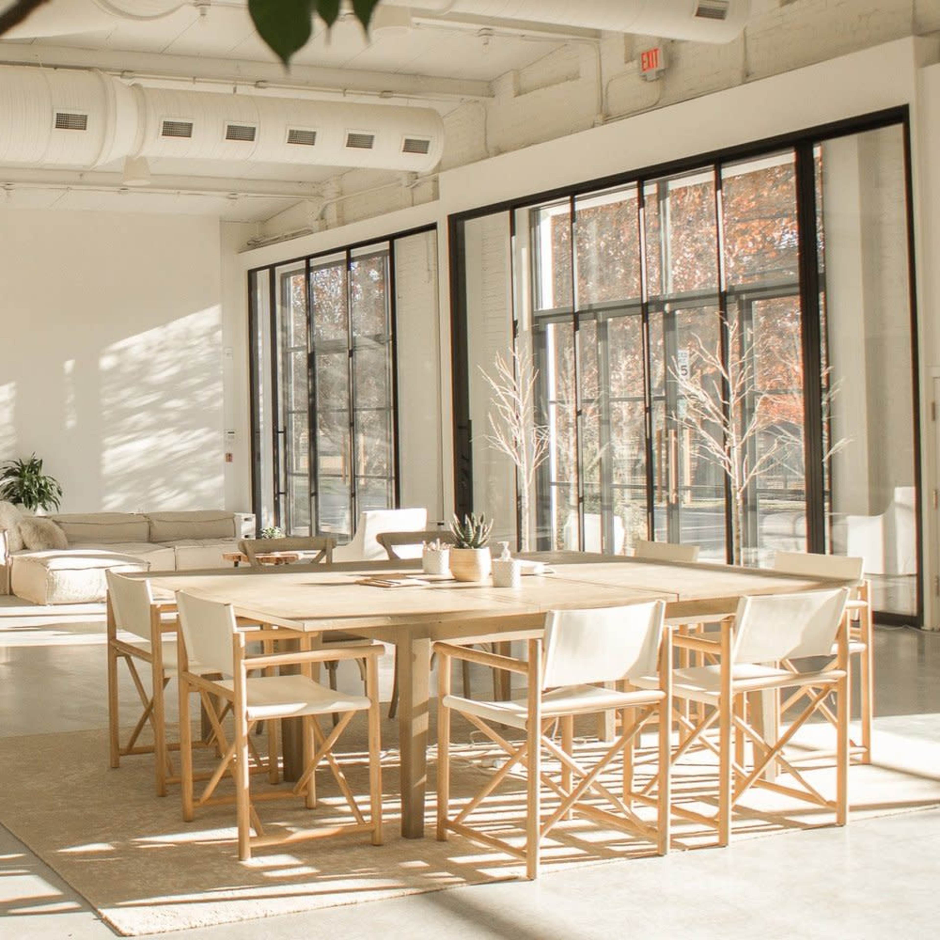 A spacious, modern interior features a large dining table with wooden chairs, a seating area, and expansive windows allowing natural light to fill the space.