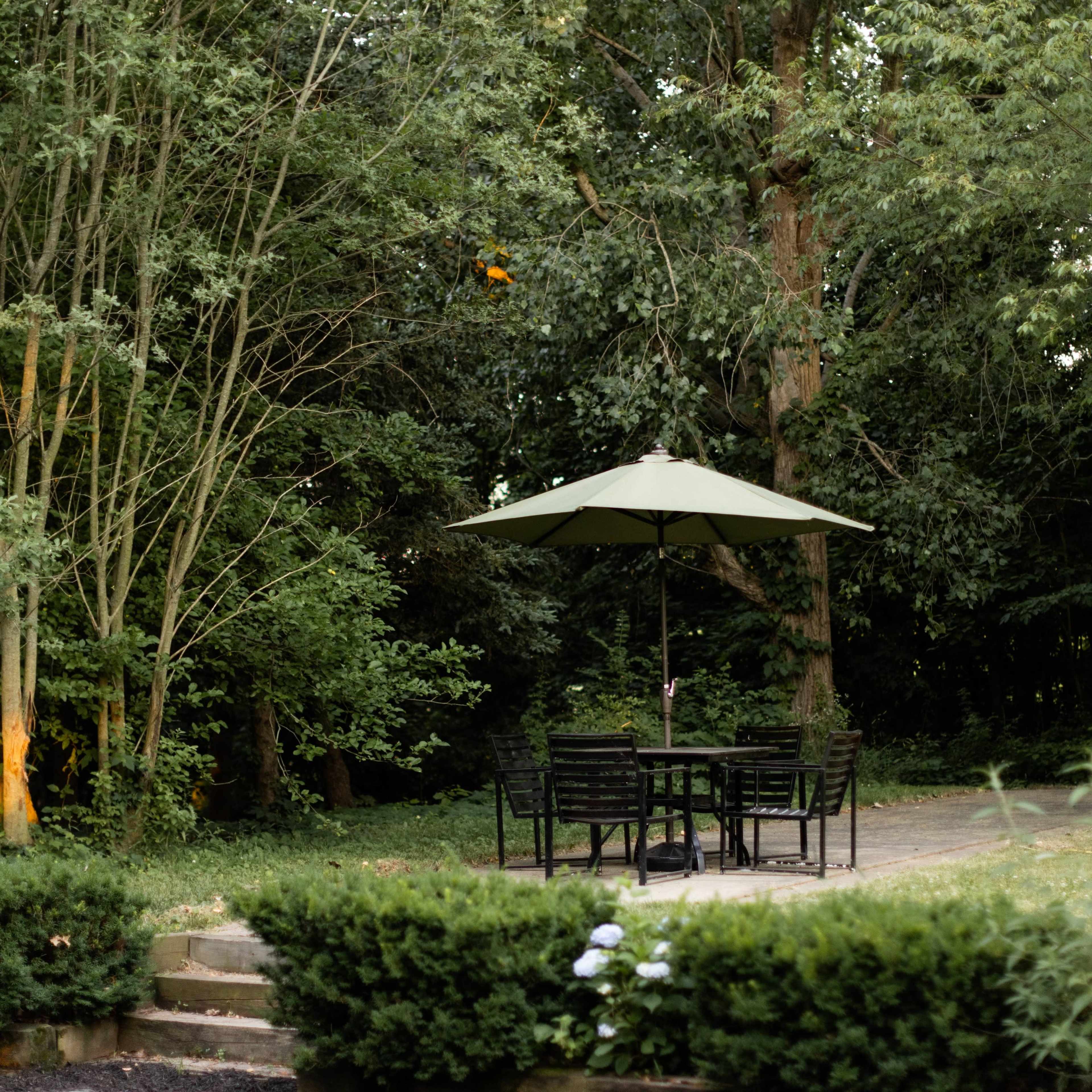 A shaded patio area with a table and chairs is set in a wooded backyard surrounded by bushes and trees.
