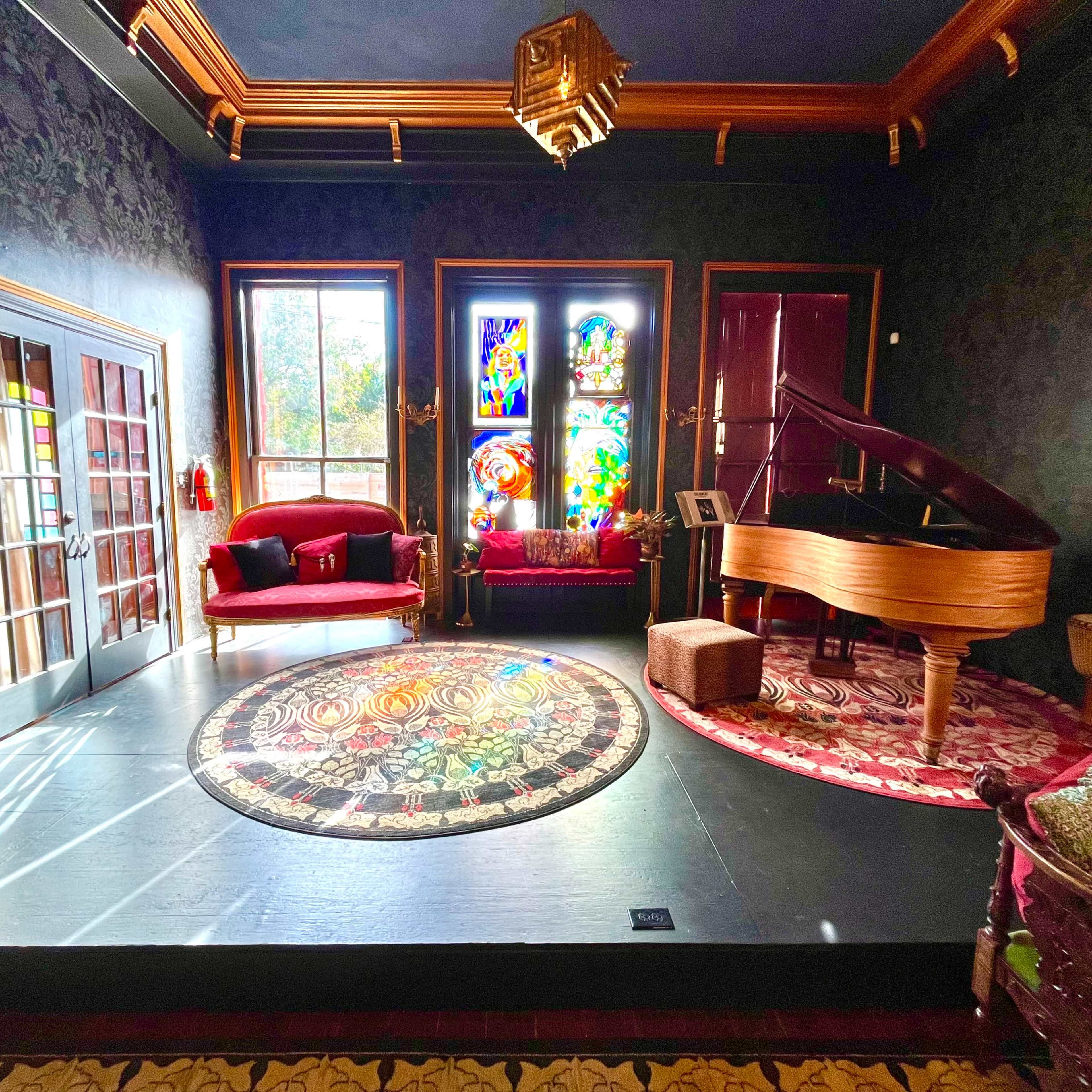 The room features a grand piano beside a colorful stained glass window, a circular rug on the floor, and a red sofa against the wall.