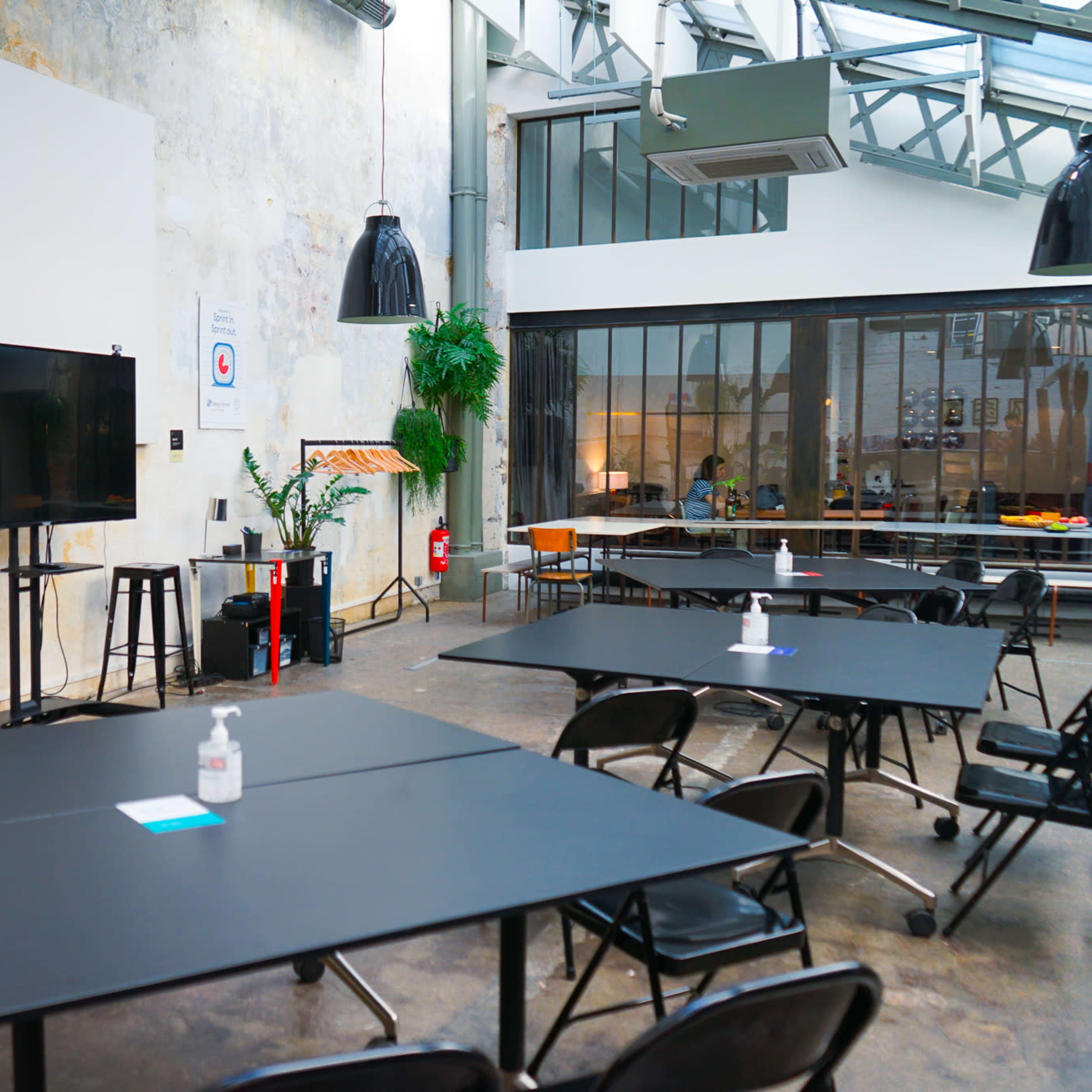 A spacious meeting room with black tables, metal stools, a large screen, and large windows allowing natural light.