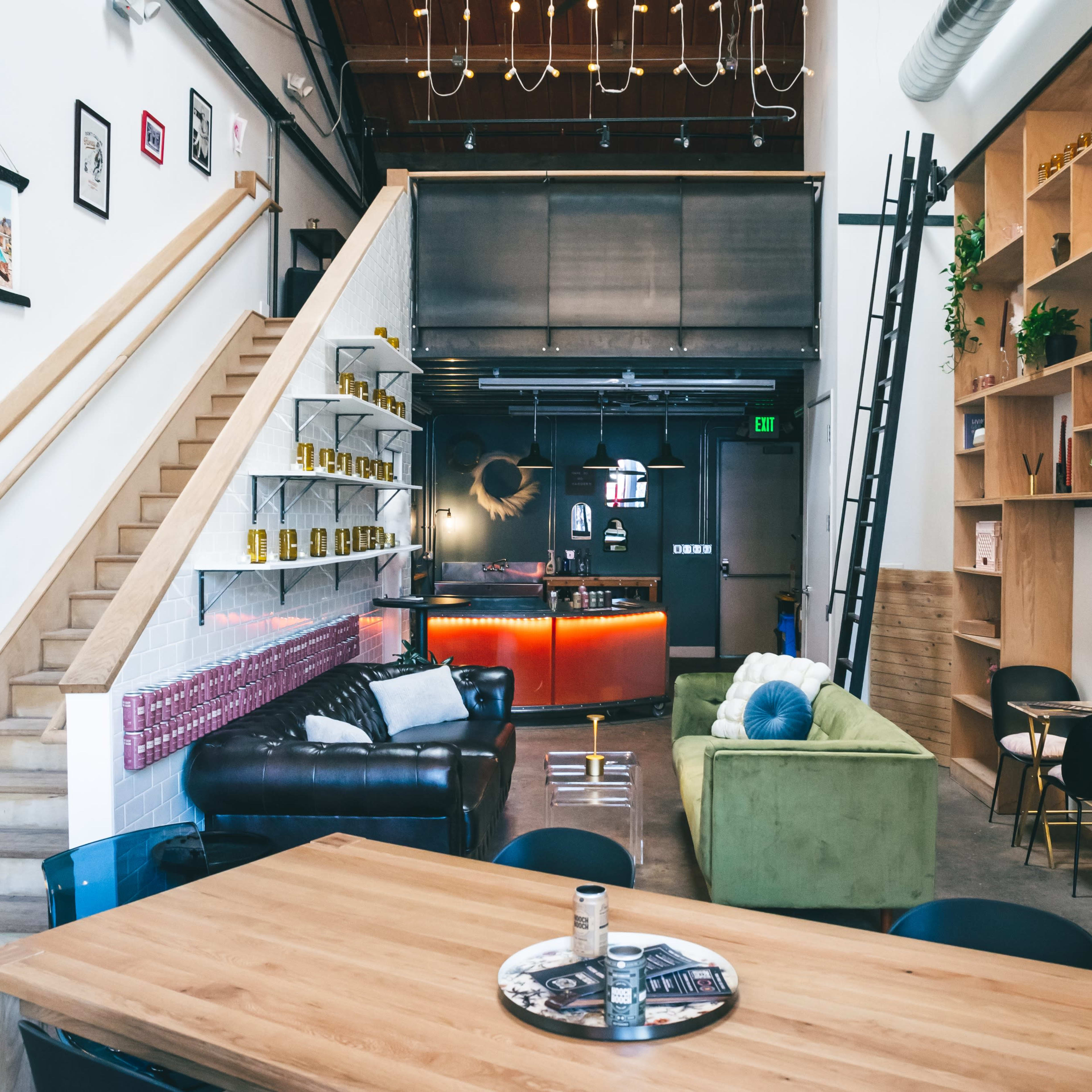 A stylish interior of a lounge area featuring a wooden dining table, green and black seating, a bar area with illuminated shelves, and a staircase leading to an upper level.