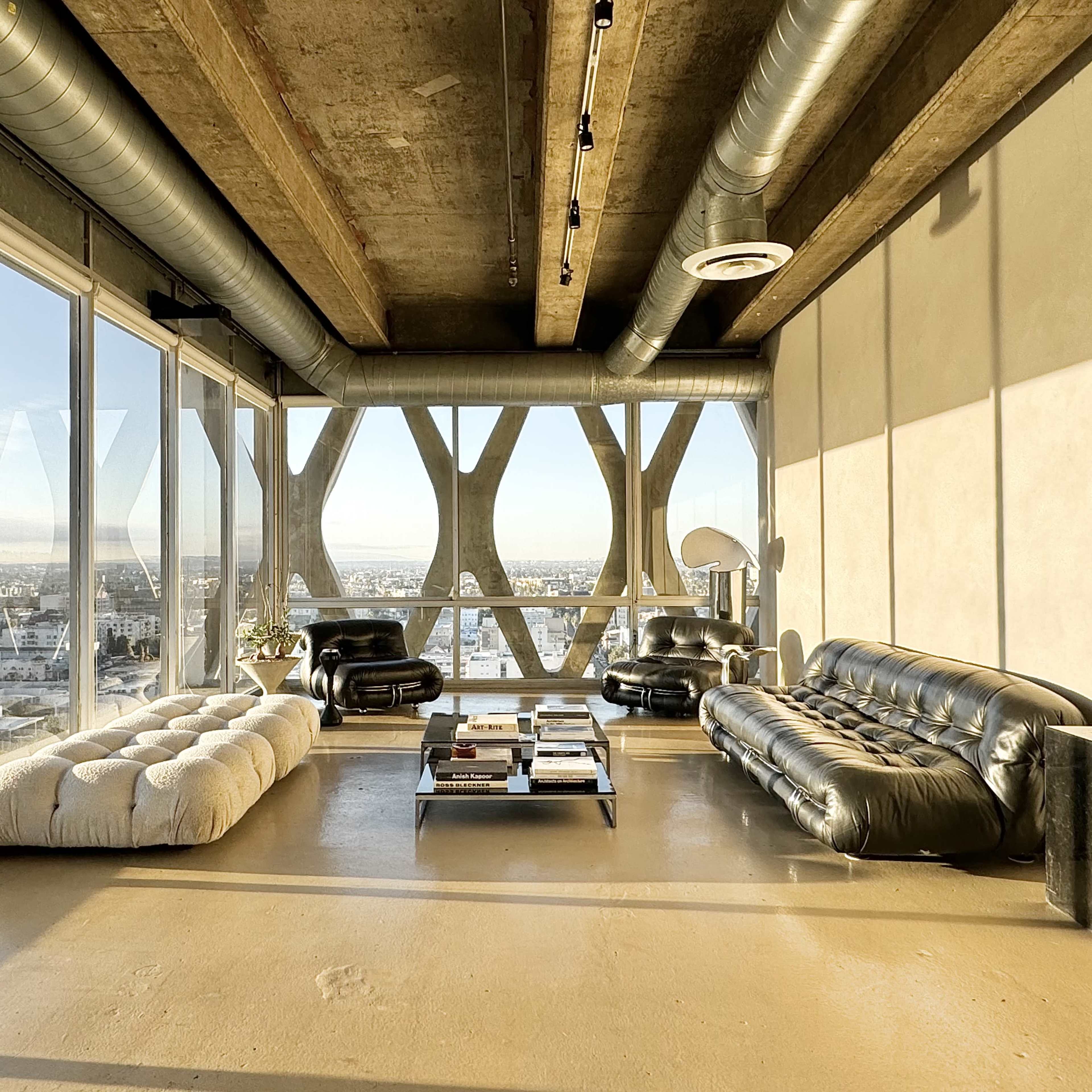 A modern living room with large windows offering a view of the city, featuring two black sofas, a coffee table, and industrial-style decor.