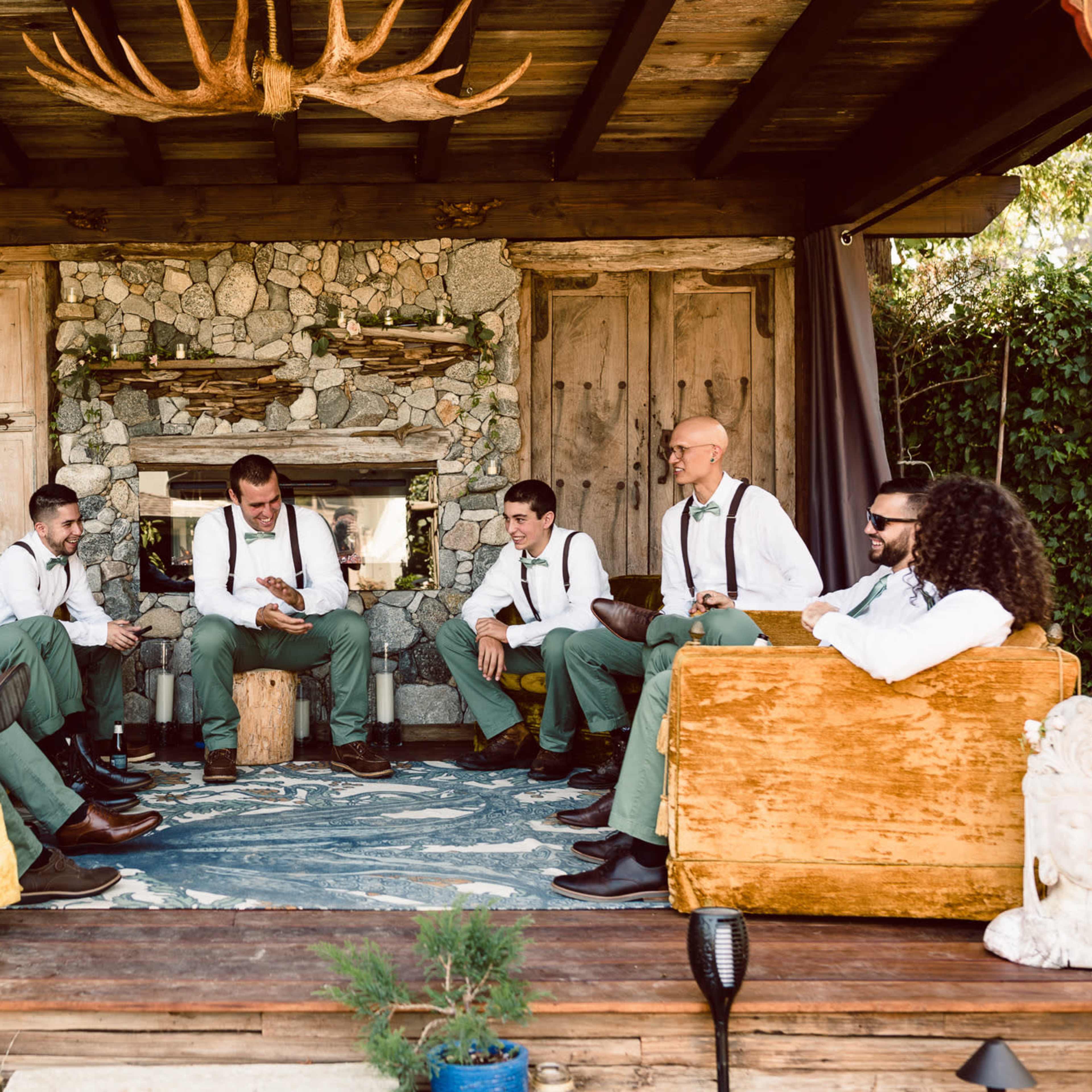 A group of men dressed in formal attire sits in a rustic setting, engaged in conversation around a wooden table in a cozy room with stone accents.