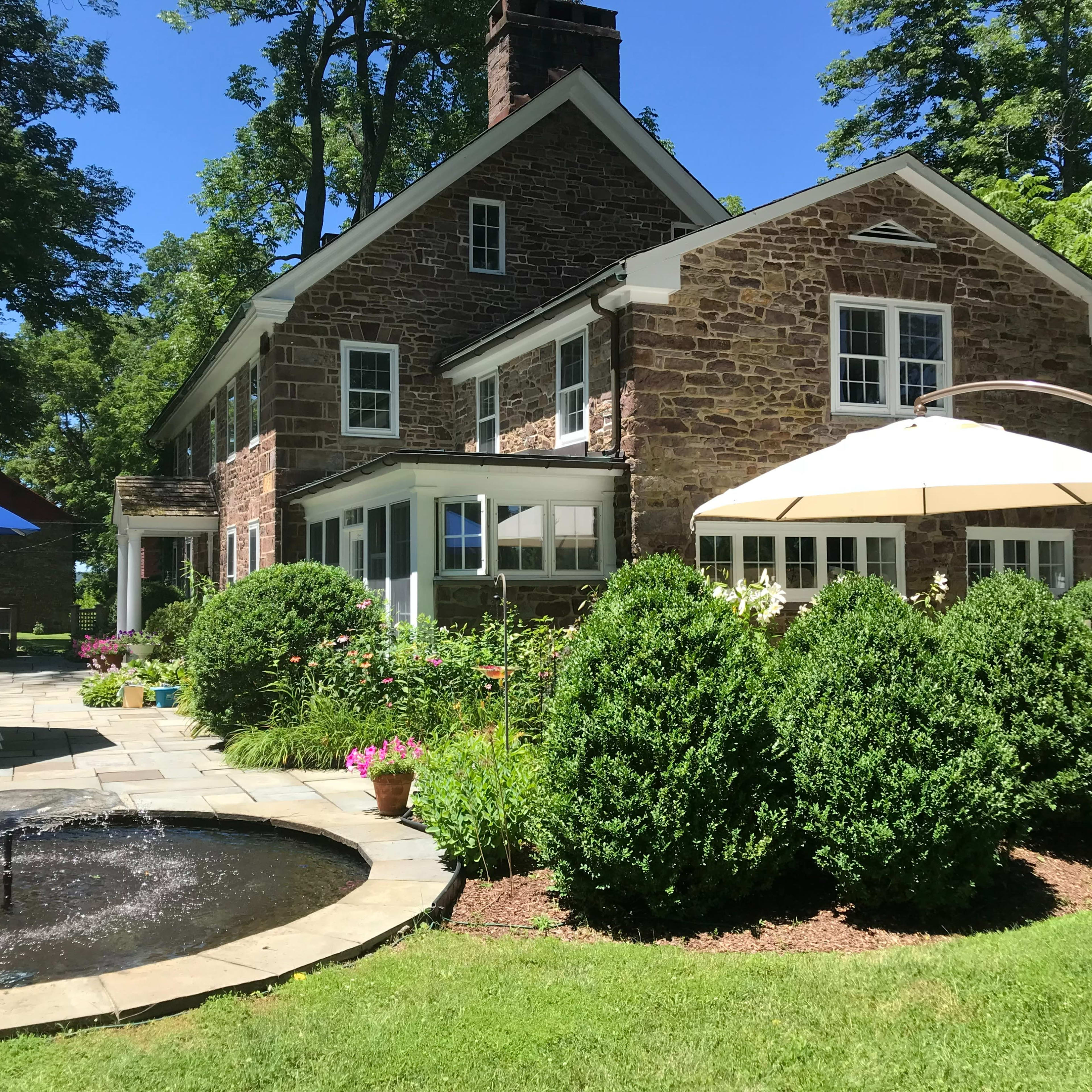 A stone house is surrounded by well-maintained gardens and a small pond with a fountain, featuring patio furniture and umbrellas for outdoor seating.