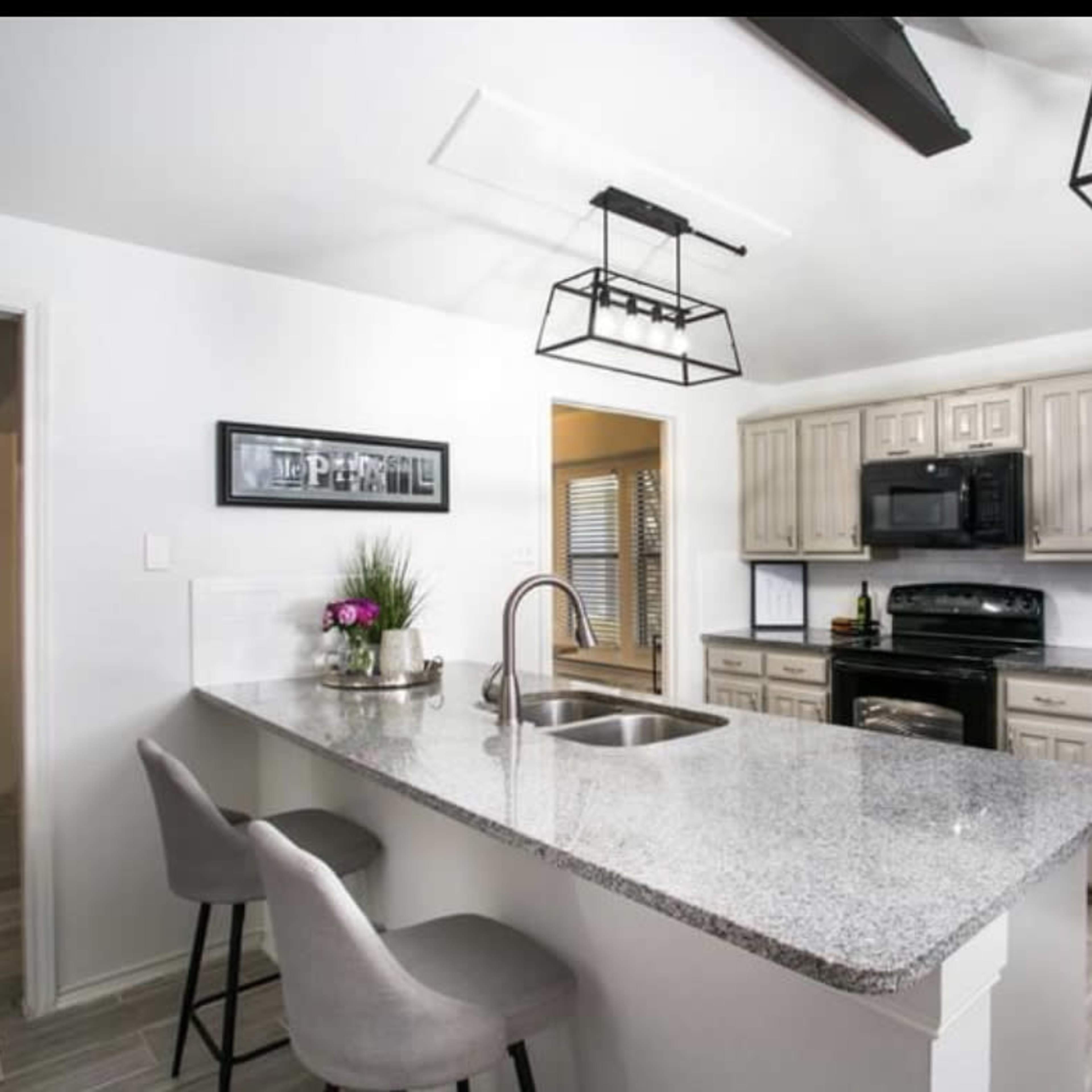 A modern kitchen with gray cabinets, black appliances, a granite countertop, and two bar stools at the island.