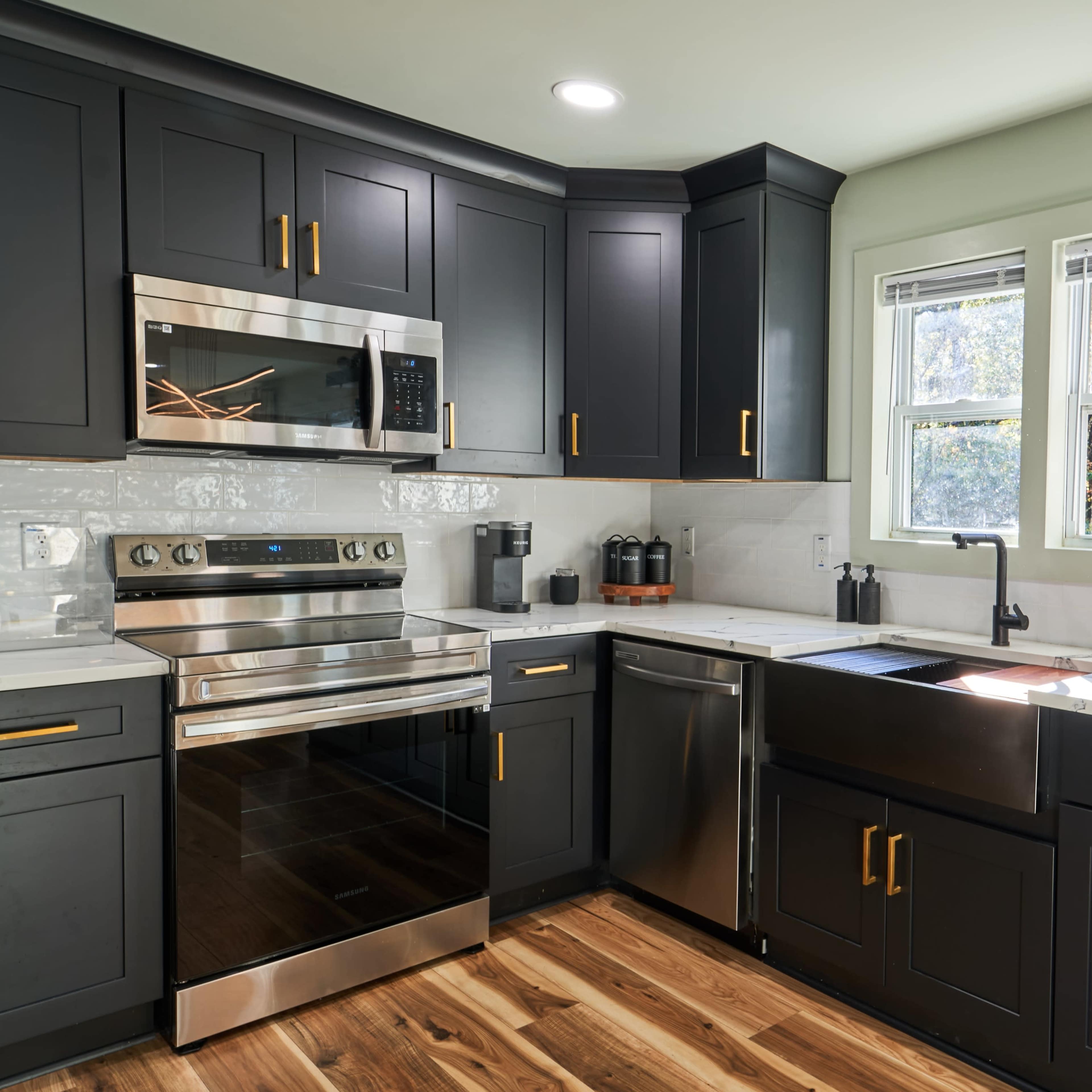 A modern kitchen with dark cabinetry, stainless steel appliances, and a marble countertop featuring a sink and window.