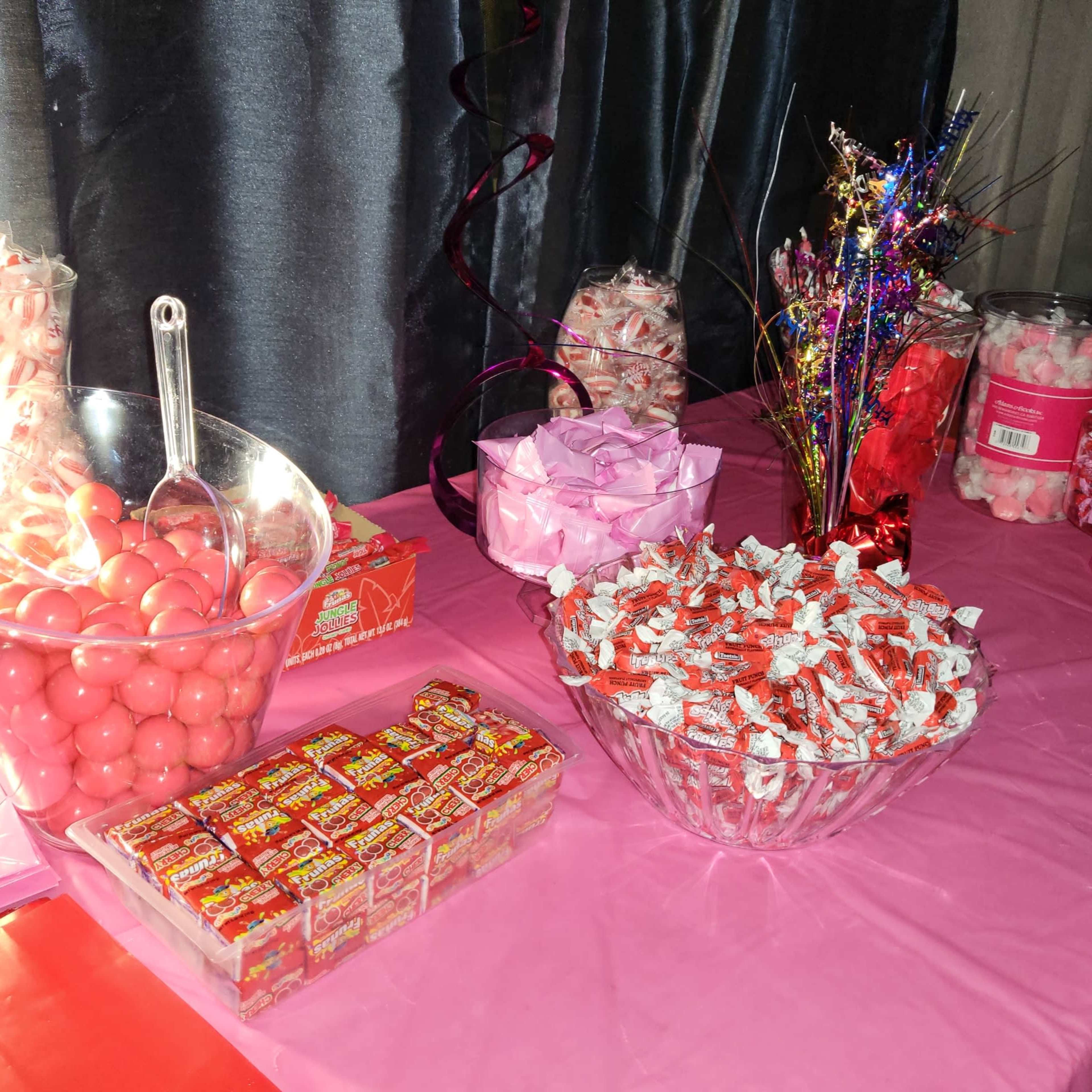 A table covered in pink cloth displays various containers filled with colorful candies, including pink gumballs, festive decorations, and assorted wrapped treats.