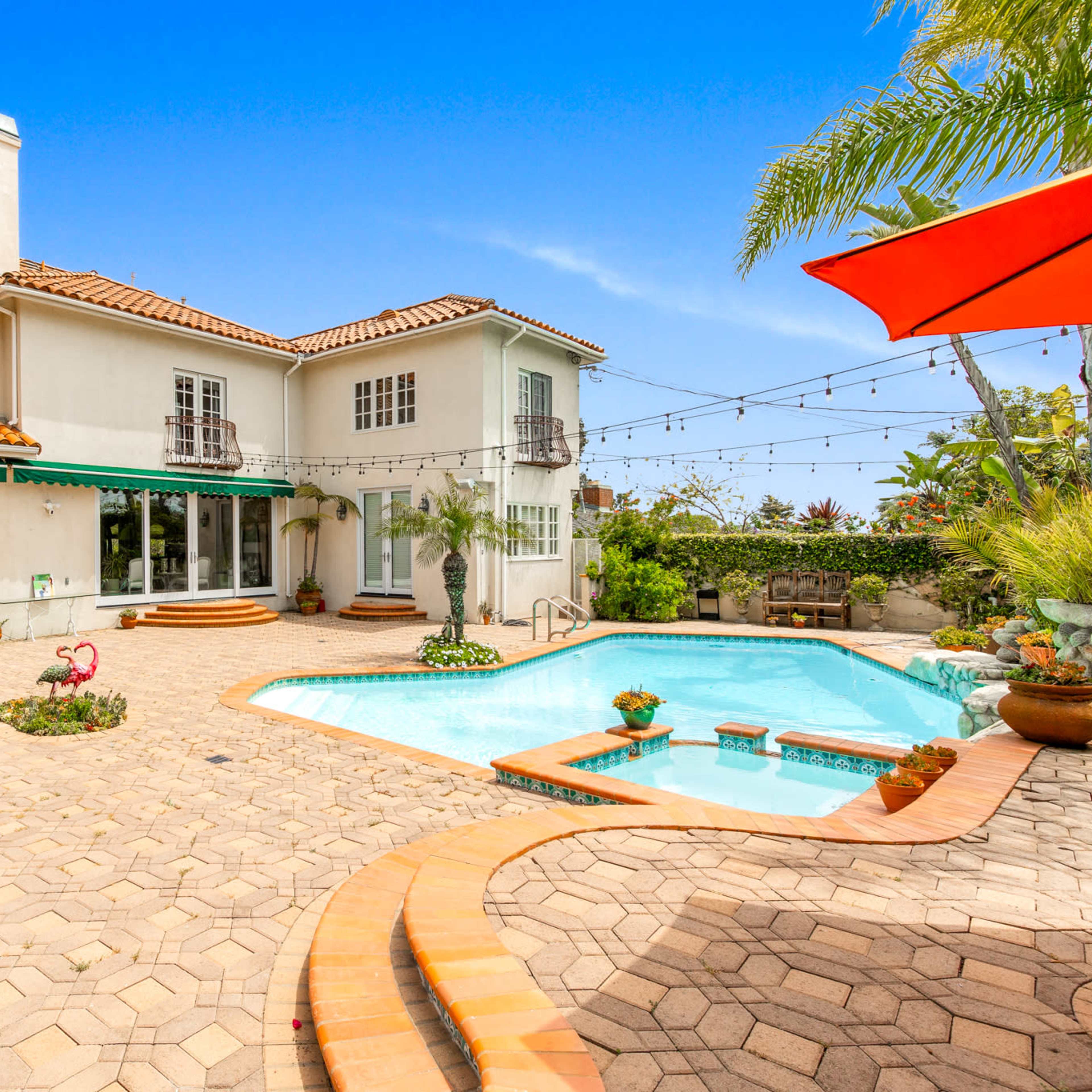 A two-story house with a red tile roof and a patio surrounds a swimming pool, flanked by potted plants and decorative features.