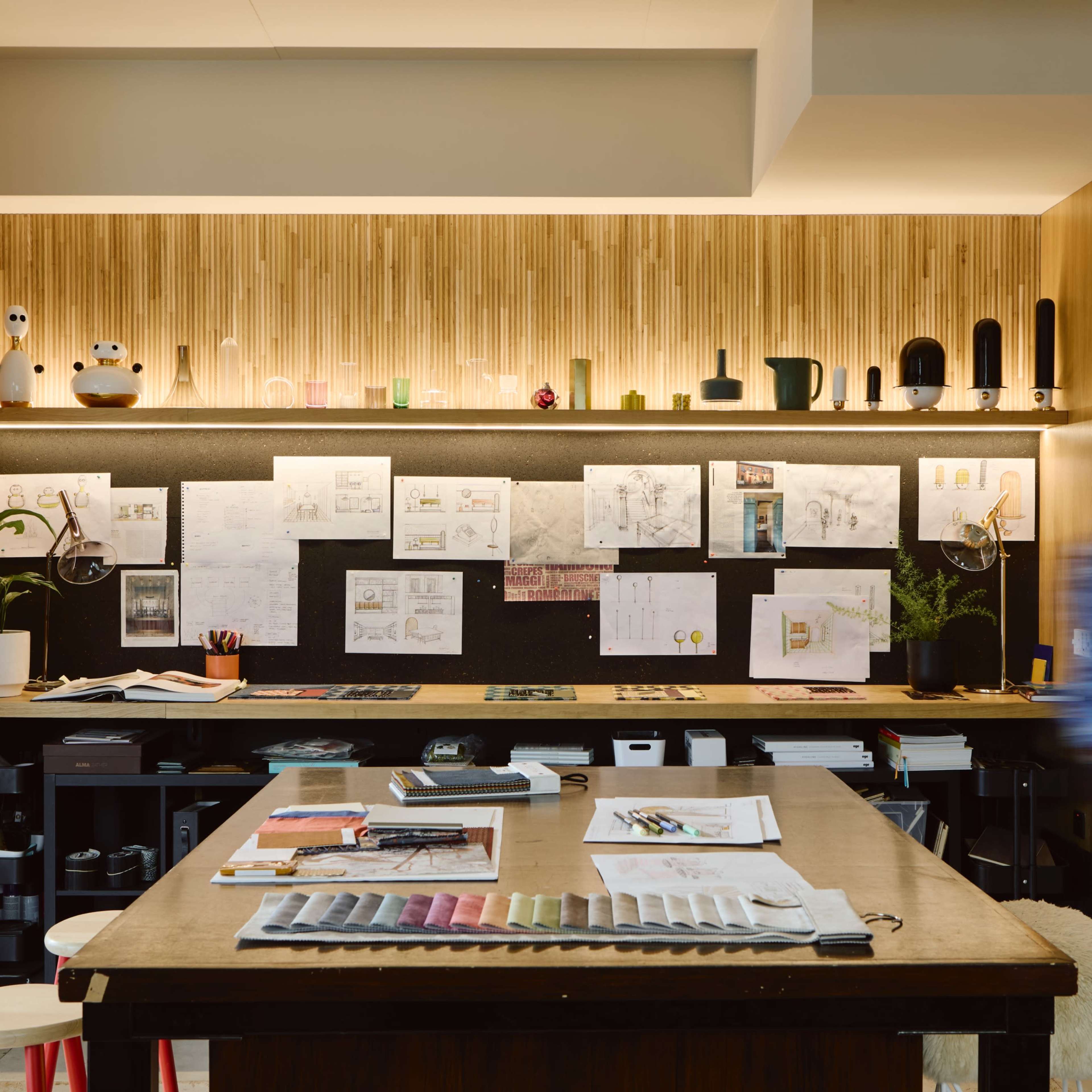 A creative workspace features a wooden table surrounded by sketches and design materials, with a wall of pinned drawings and a softly lit shelf showcasing various objects.