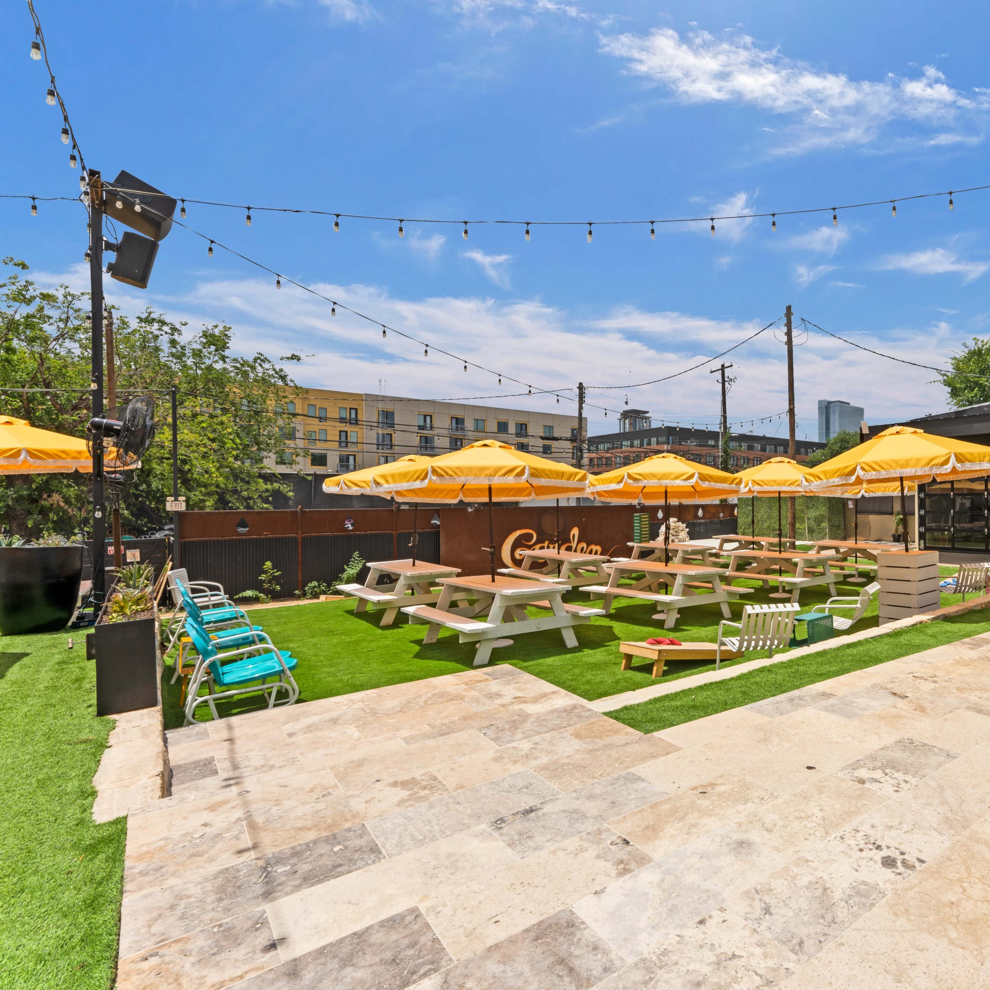 A vibrant outdoor dining area with picnic tables, umbrellas, and string lights against a backdrop of greenery and a modern building.