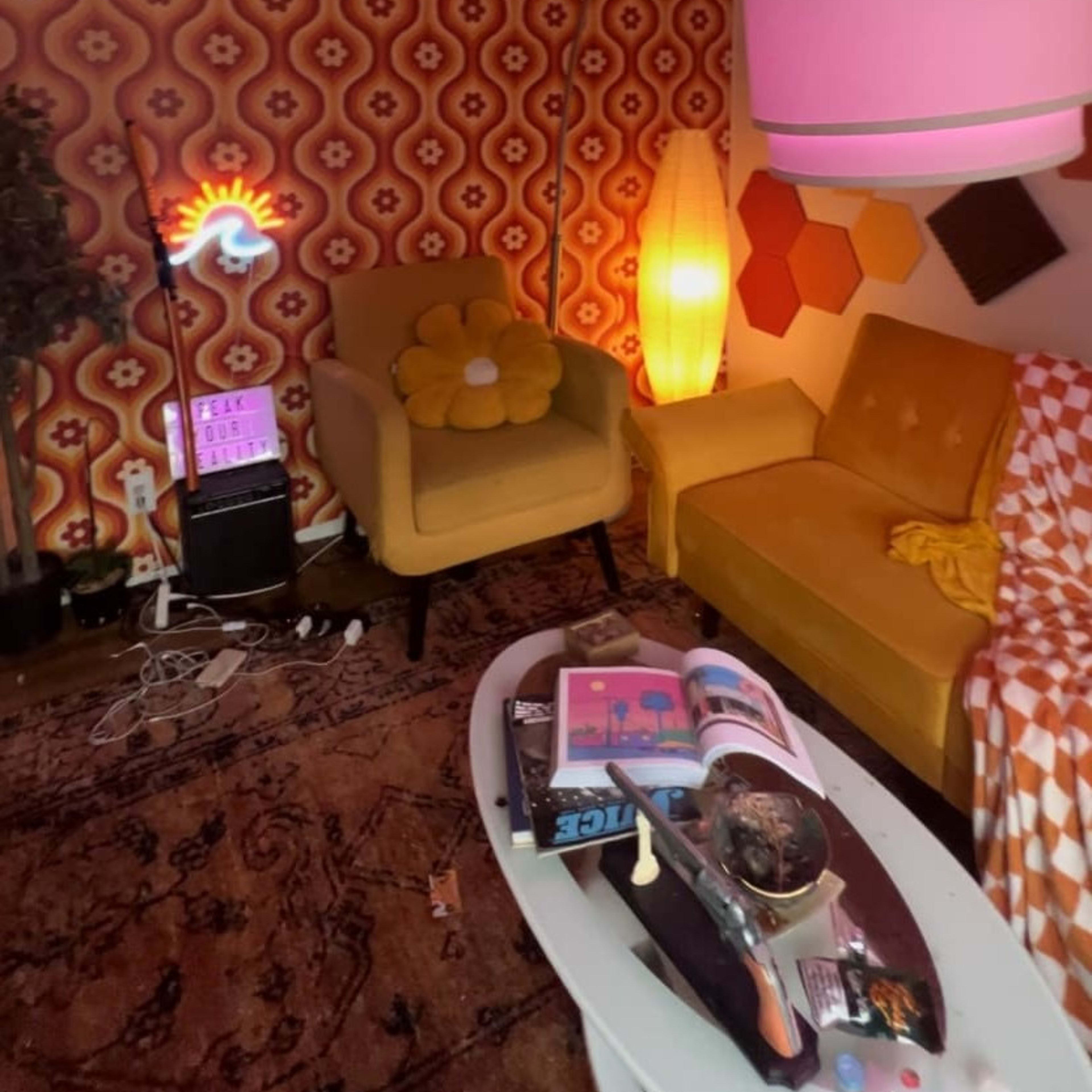 A retro-styled living room with patterned wallpaper, a yellow sofa, an armchair with a flower design, a coffee table cluttered with books and decorative items, and various lighting sources.