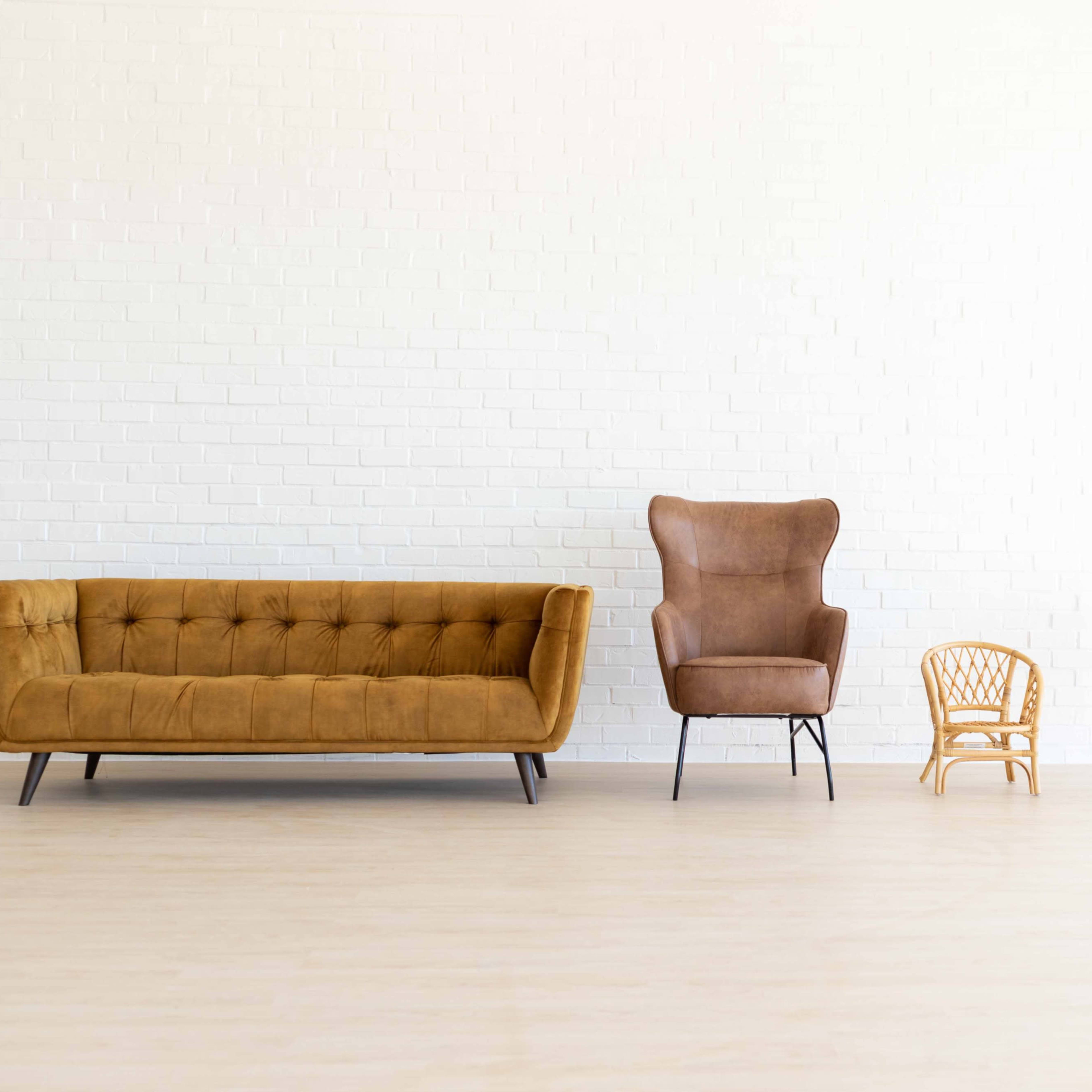 A minimalist interior featuring a brown velvet sofa, a brown upholstered armchair, a small wooden chair, and a white circular chair against a white brick wall.